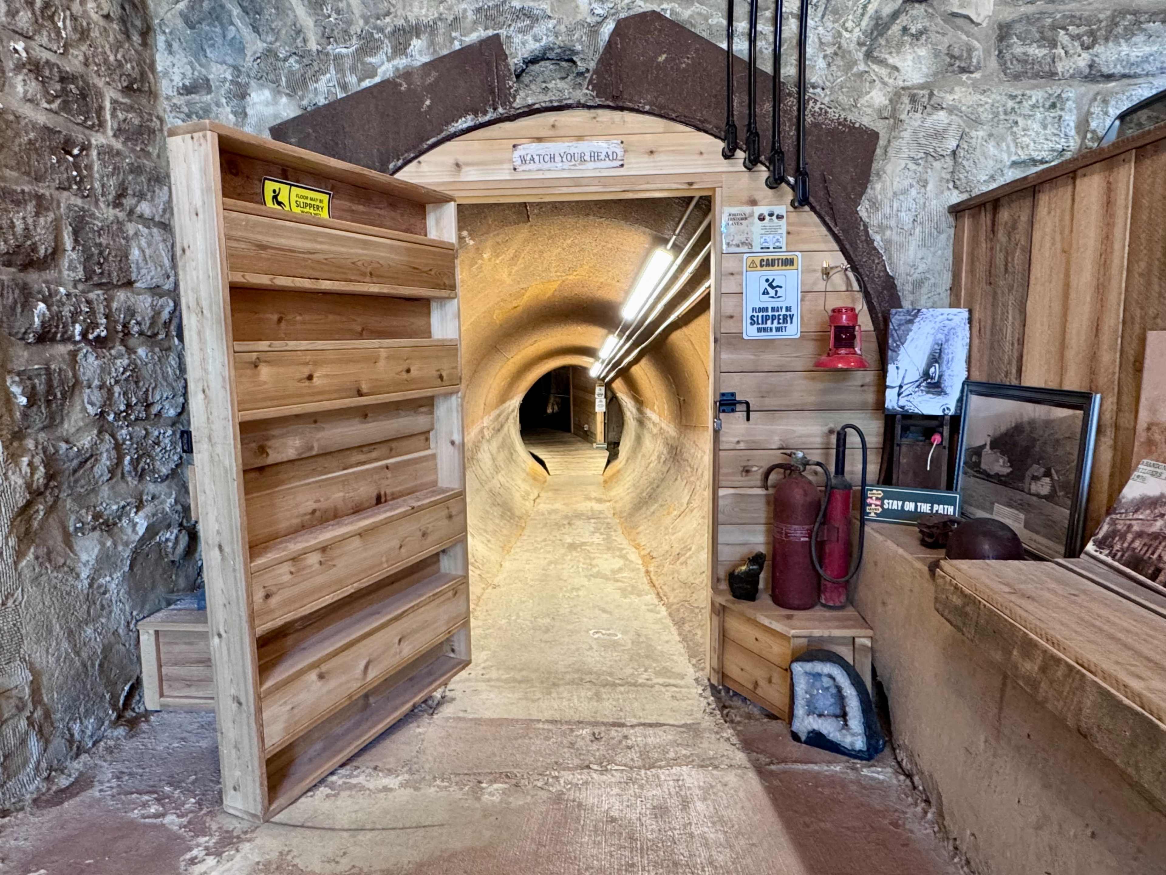 The image shows a circular tunnel entrance with wooden doors, lined with lights and stone walls, leading into a dimly lit passage.