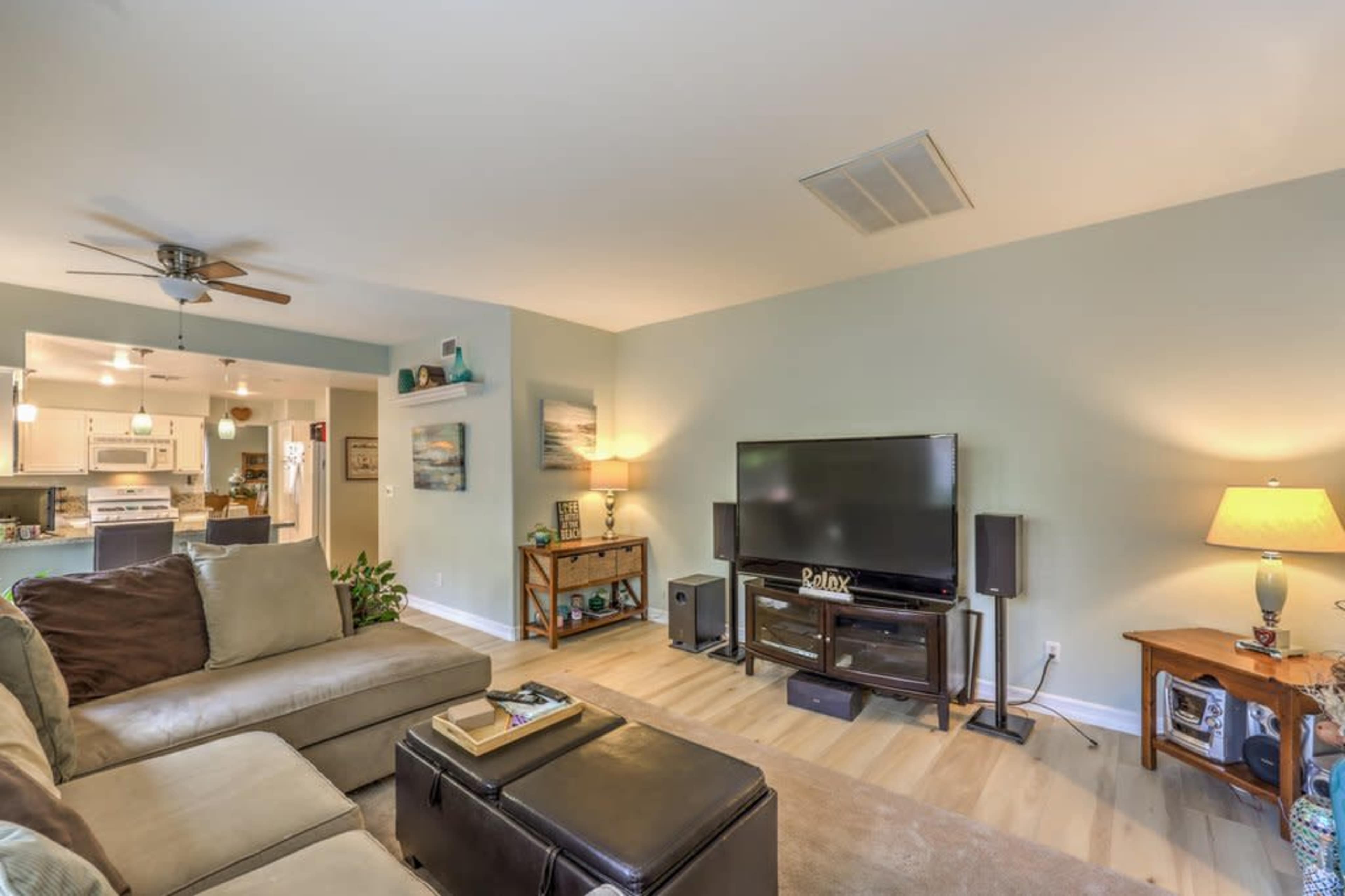 A living room featuring a sectional sofa, a large flat-screen TV on a stand, and a wooden coffee table, with a light color scheme and wooden flooring.