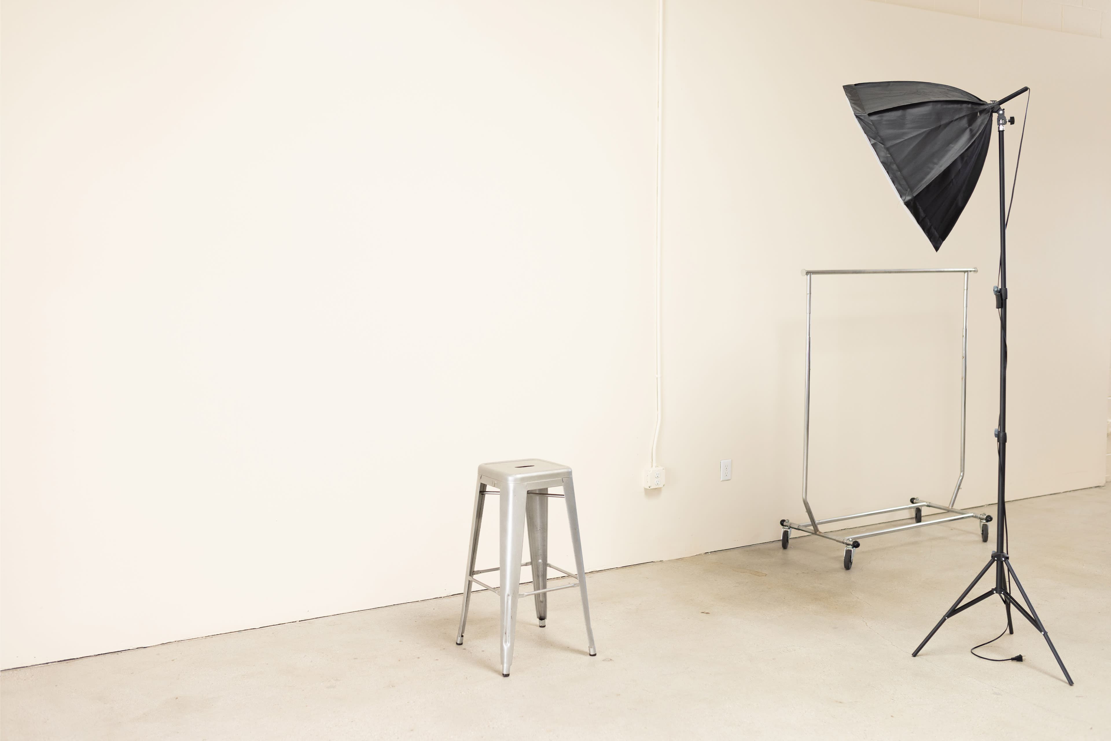 The image shows a minimalist studio space with a silver stool, a clothing rack, and a black softbox light set up against a plain white wall.