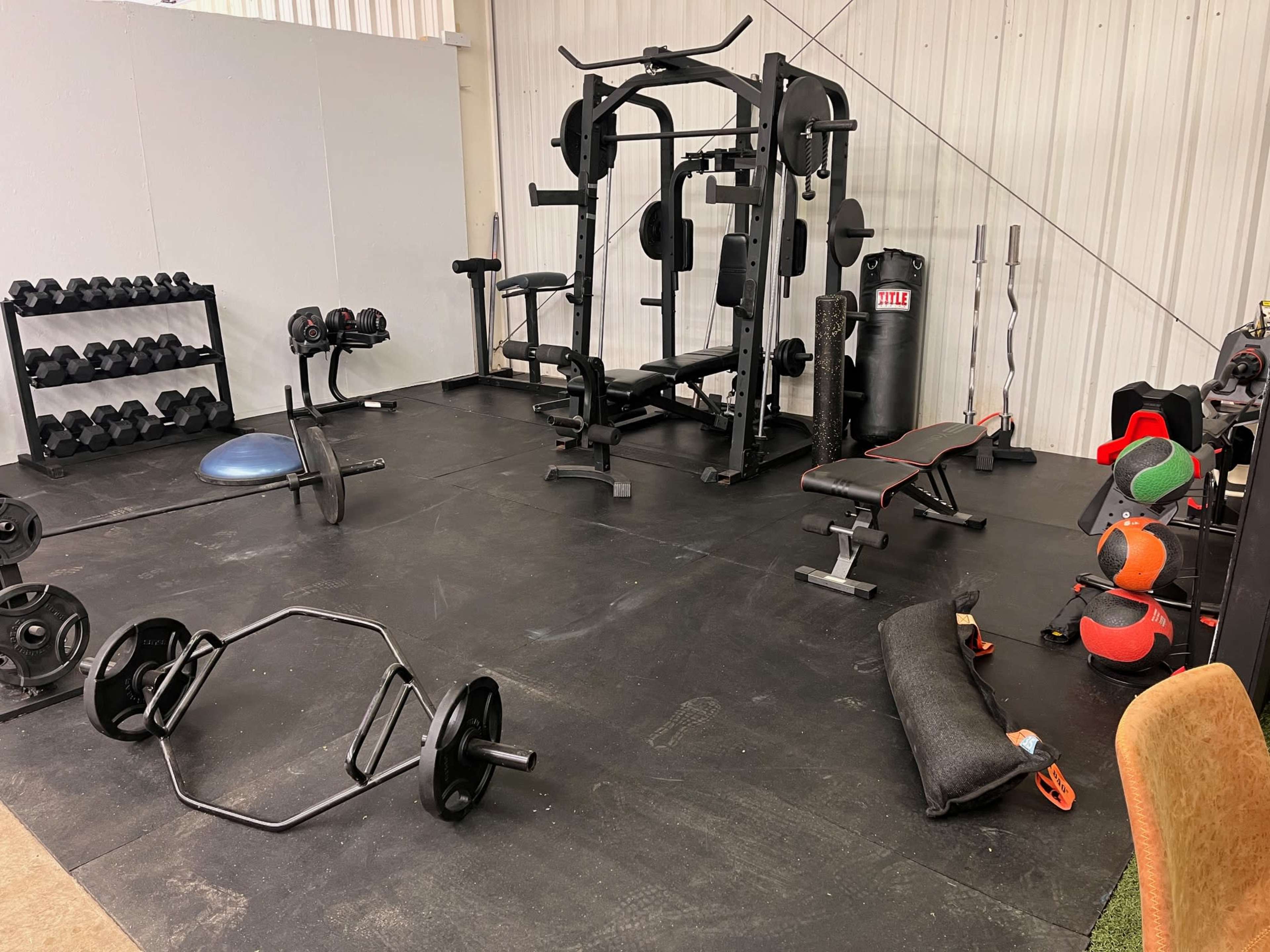 The image shows a well-equipped gym with various weights, a squat rack, benches, and exercise equipment laid out on a rubber flooring surface.