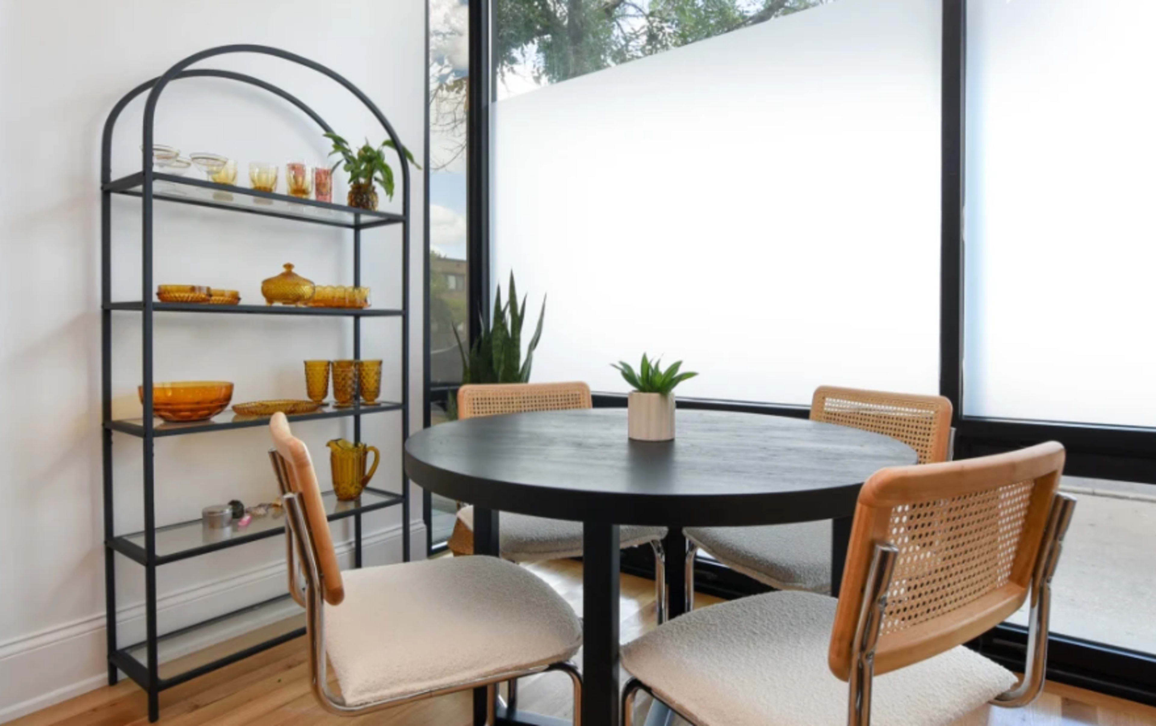 A black round table is surrounded by four chairs in a well-lit room featuring a tall shelving unit with glassware and a potted plant.