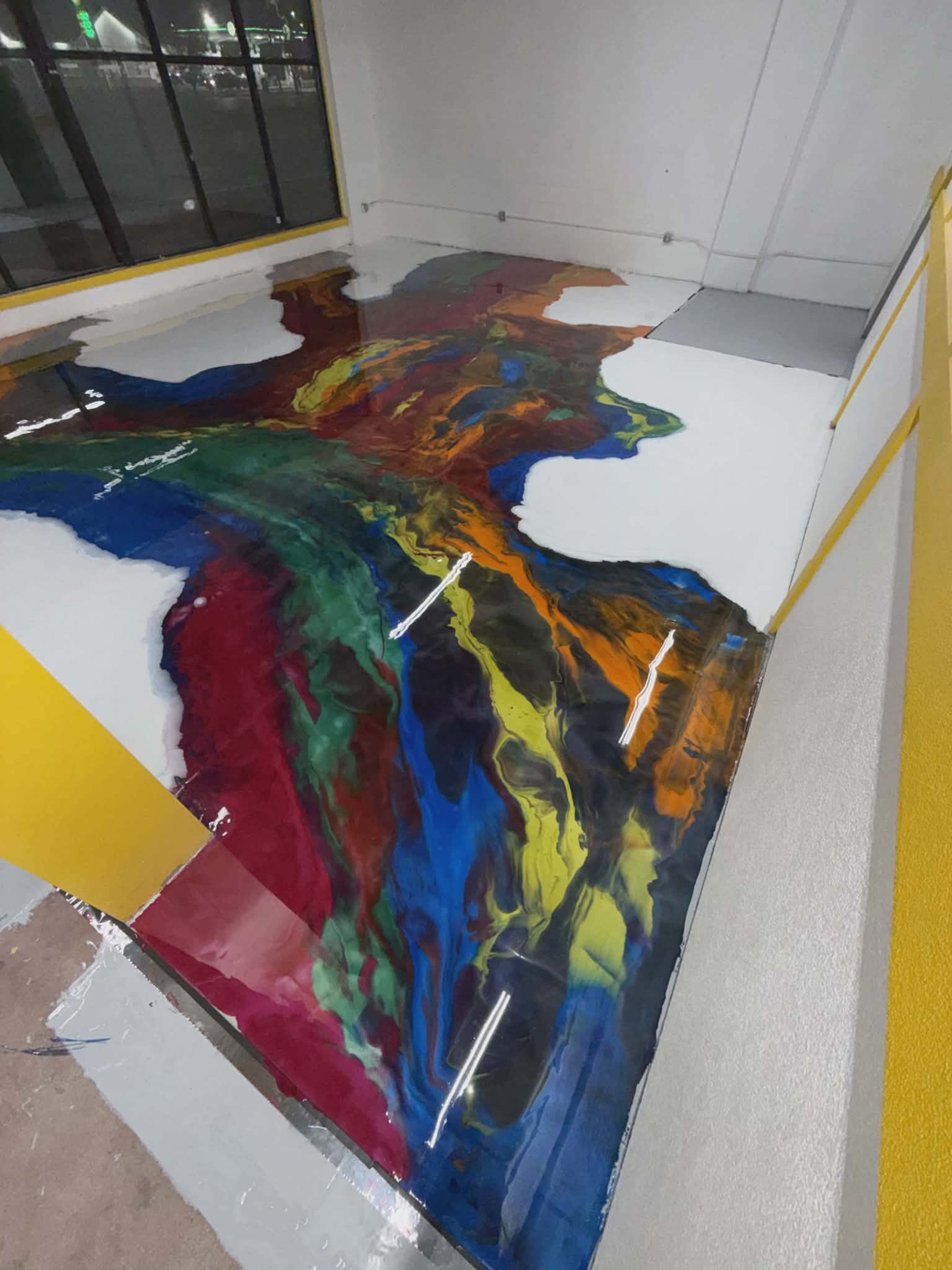 The image shows a floor with a colorful, swirling epoxy resin design that resembles a river flowing across a white surface.