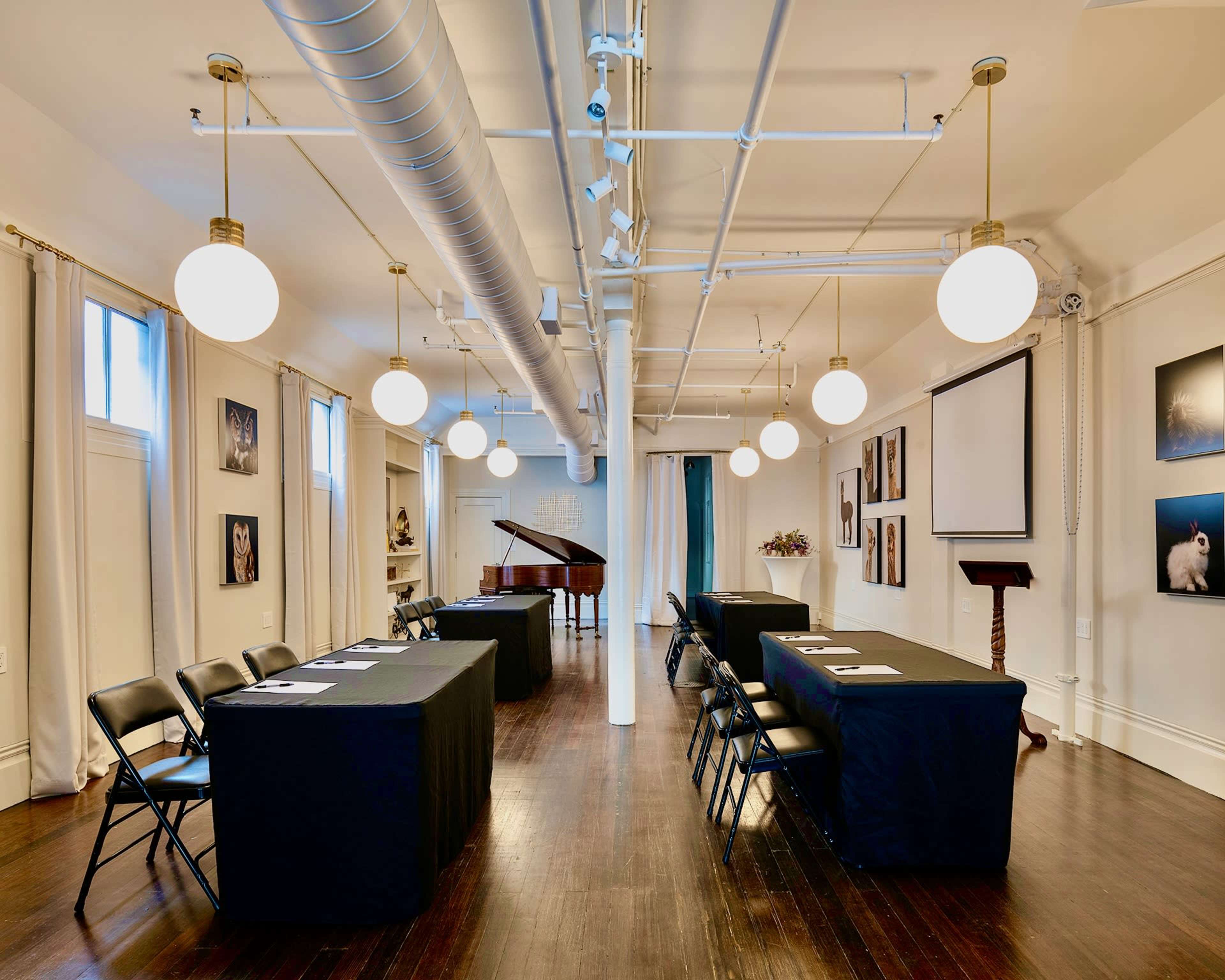 A well-lit conference room features two rows of tables covered in black cloth, with chairs arranged on either side and a grand piano in the corner.