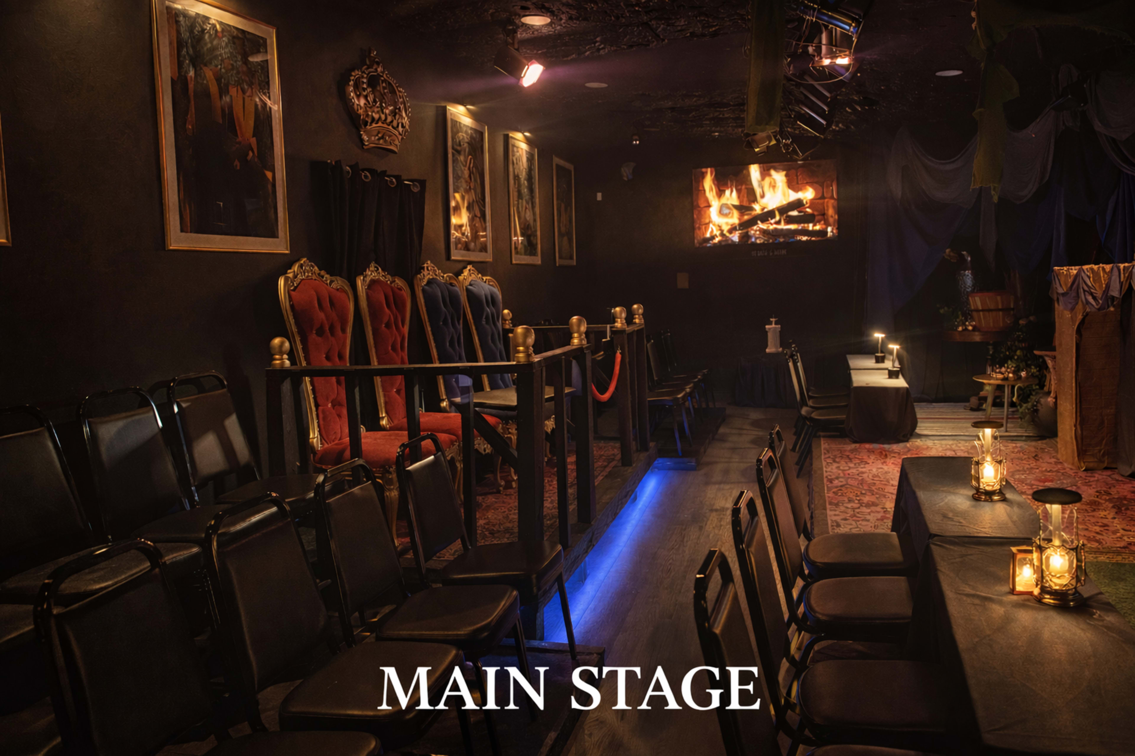 The image depicts a dimly lit theater interior featuring a main stage with red and gold throne-like chairs, a fireplace above the stage, and tables with lanterns for guests.