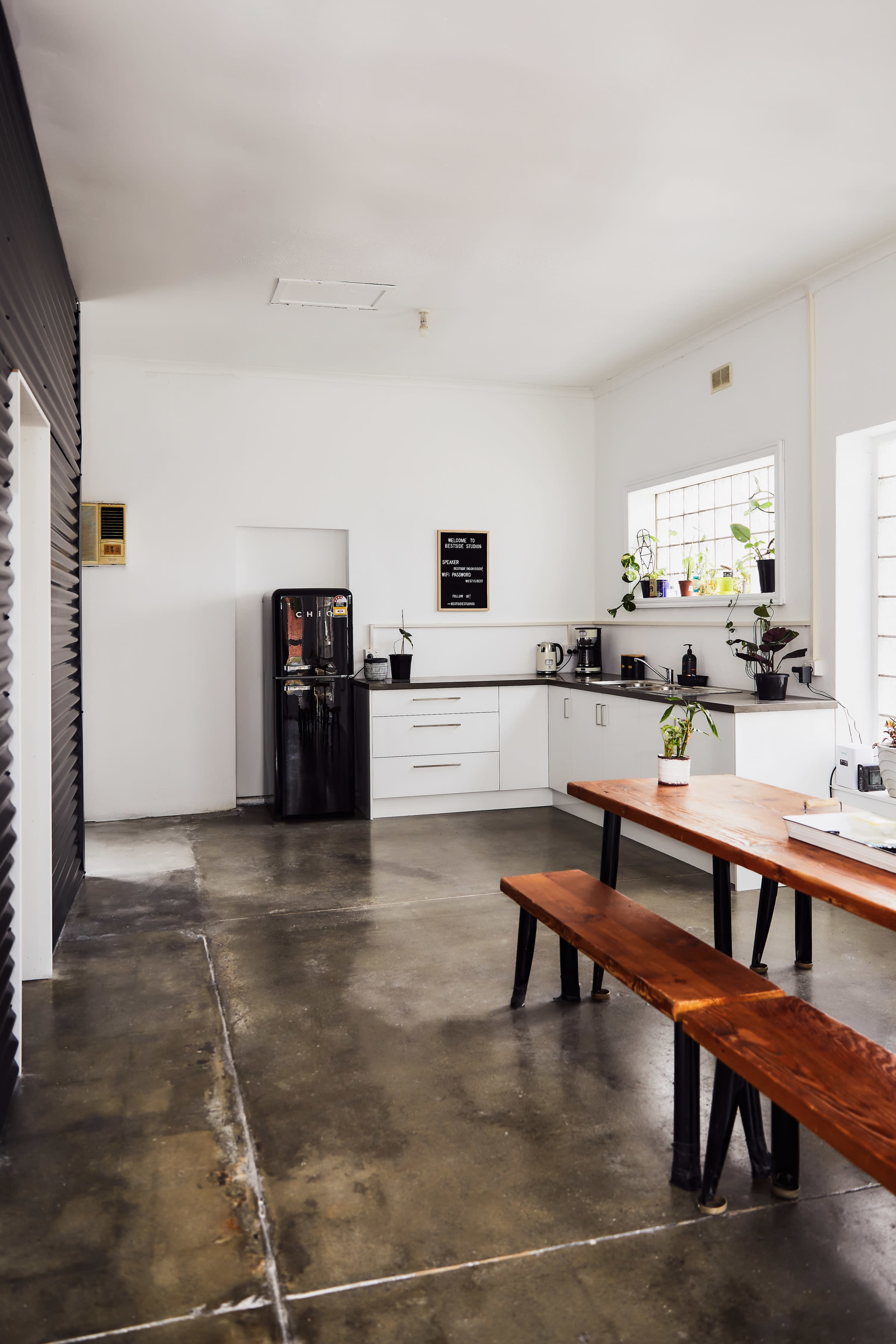 A minimalist kitchen features a black refrigerator, white cabinets, and a wooden dining table with benches.