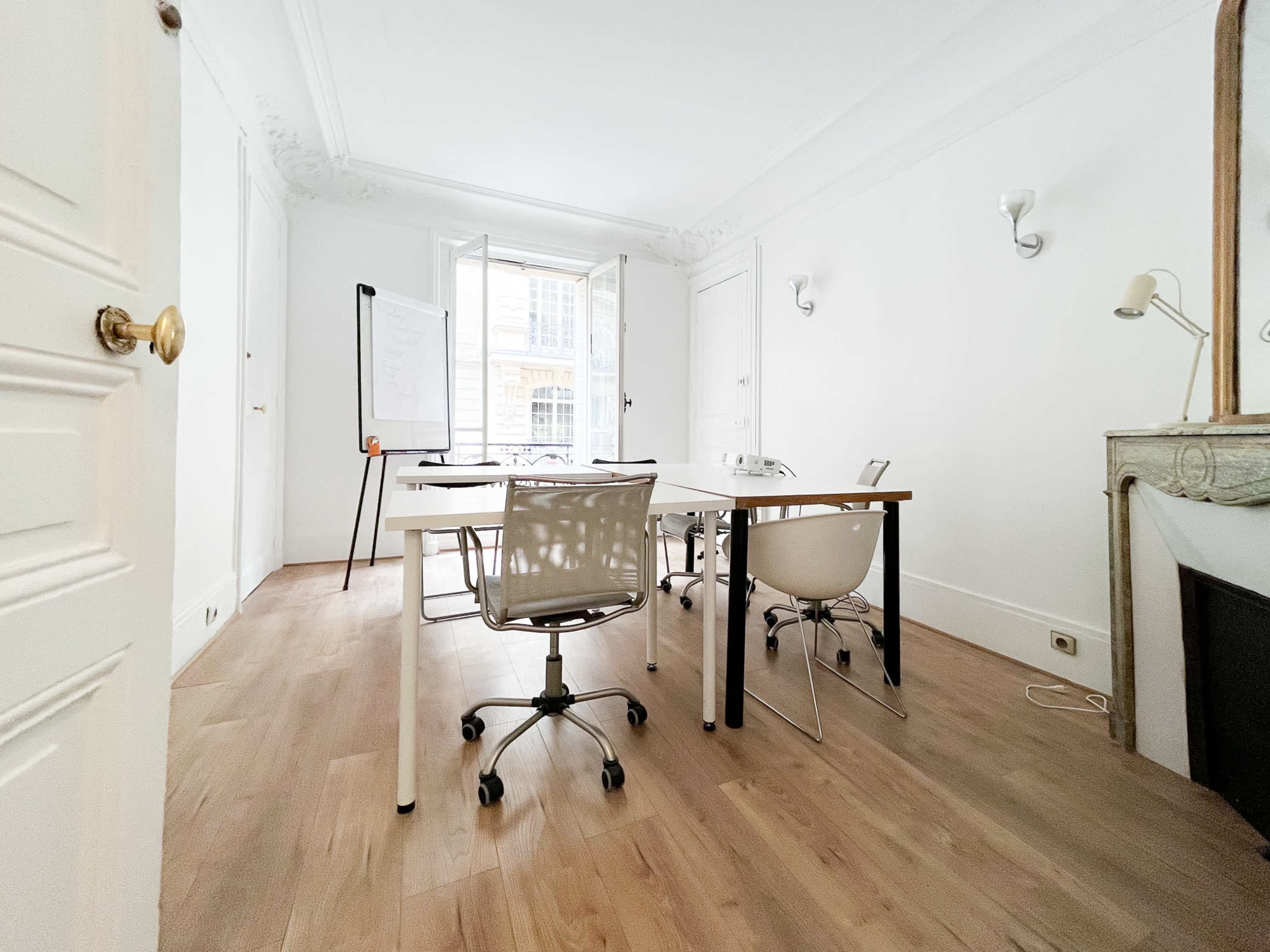 A bright, minimalist office space with a large window, a whiteboard on a stand, and two desks with chairs arranged around them.
