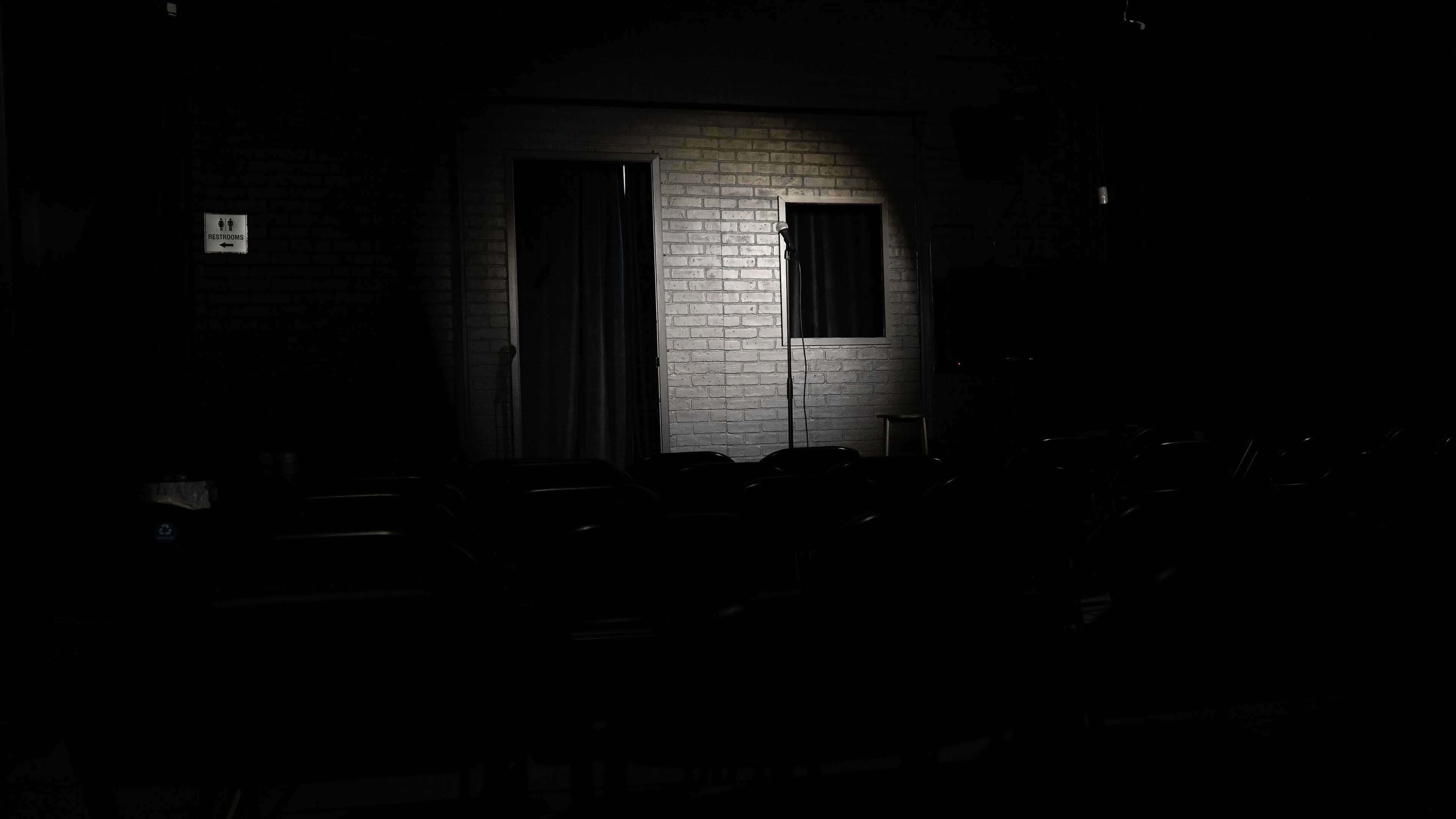 A spotlight illuminates a microphone stand in an empty stage area, framed by dark seating and a closed door.