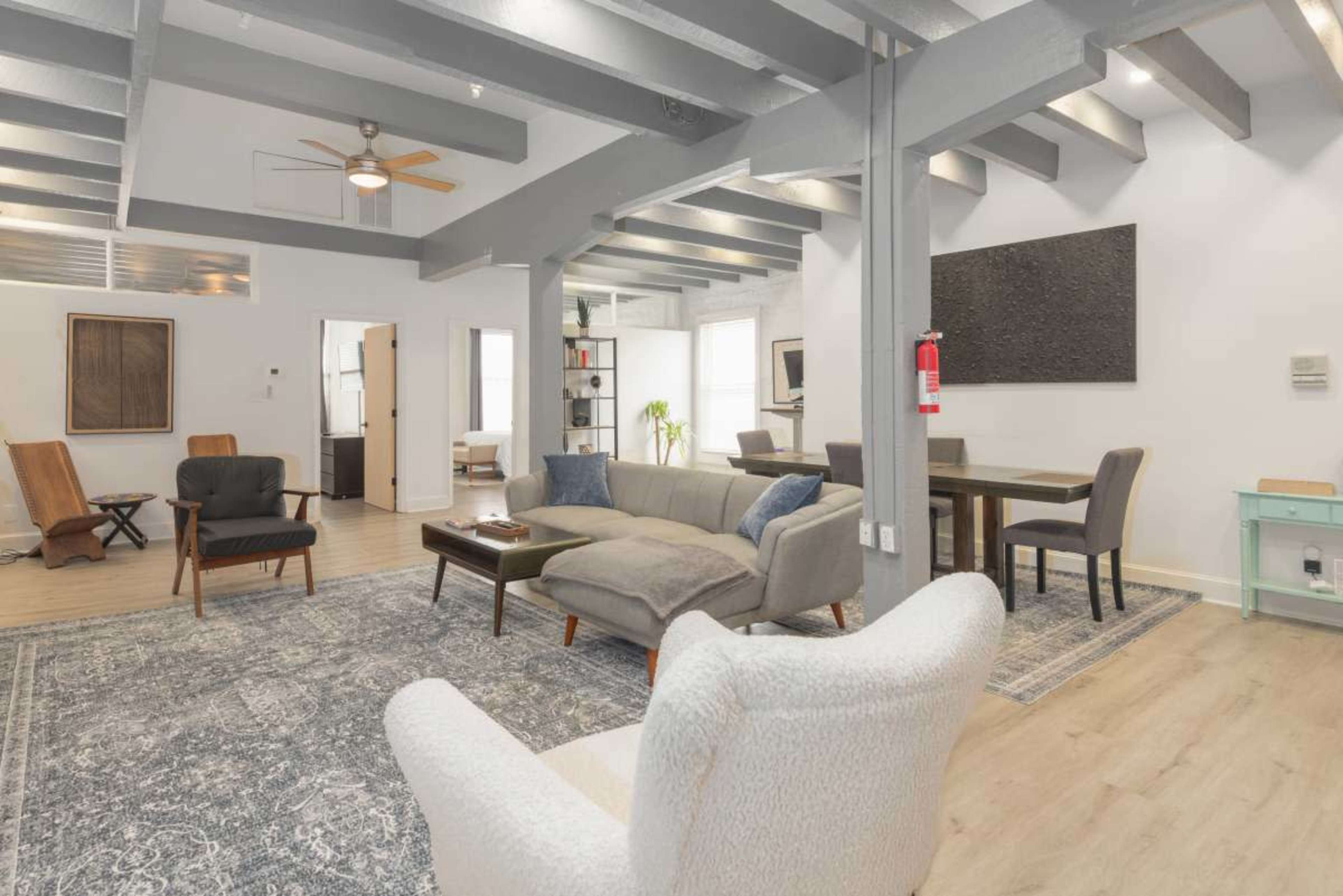 The image shows a spacious living area with a mix of seating options, wooden beams on the ceiling, and a dining table, all arranged on a textured rug.