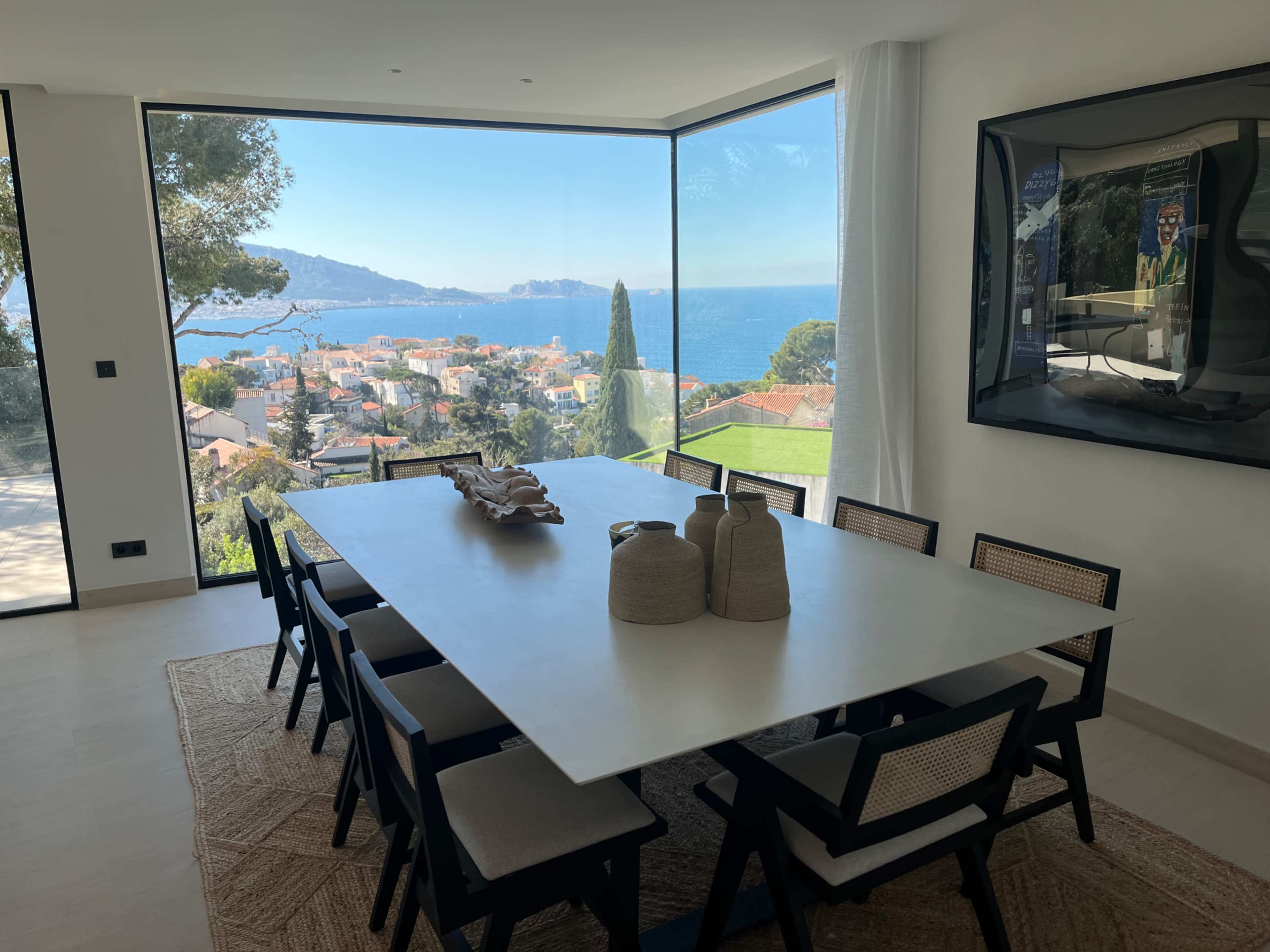 A dining room features a large white table surrounded by black chairs, overlooking a coastal view through large glass windows.