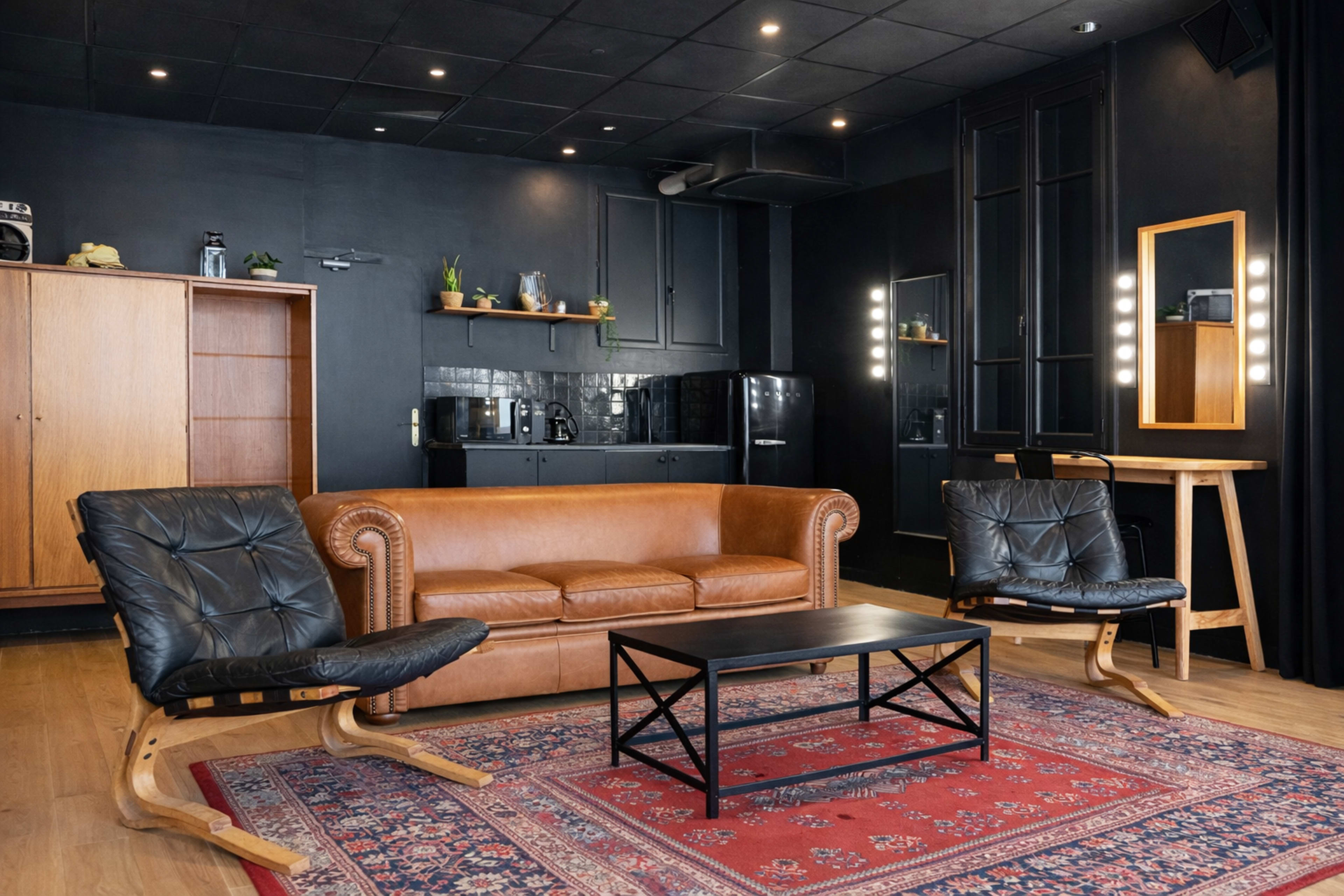 The image shows a stylish living space featuring a brown leather sofa, two black chairs, a coffee table on a patterned rug, and a kitchenette in the background.