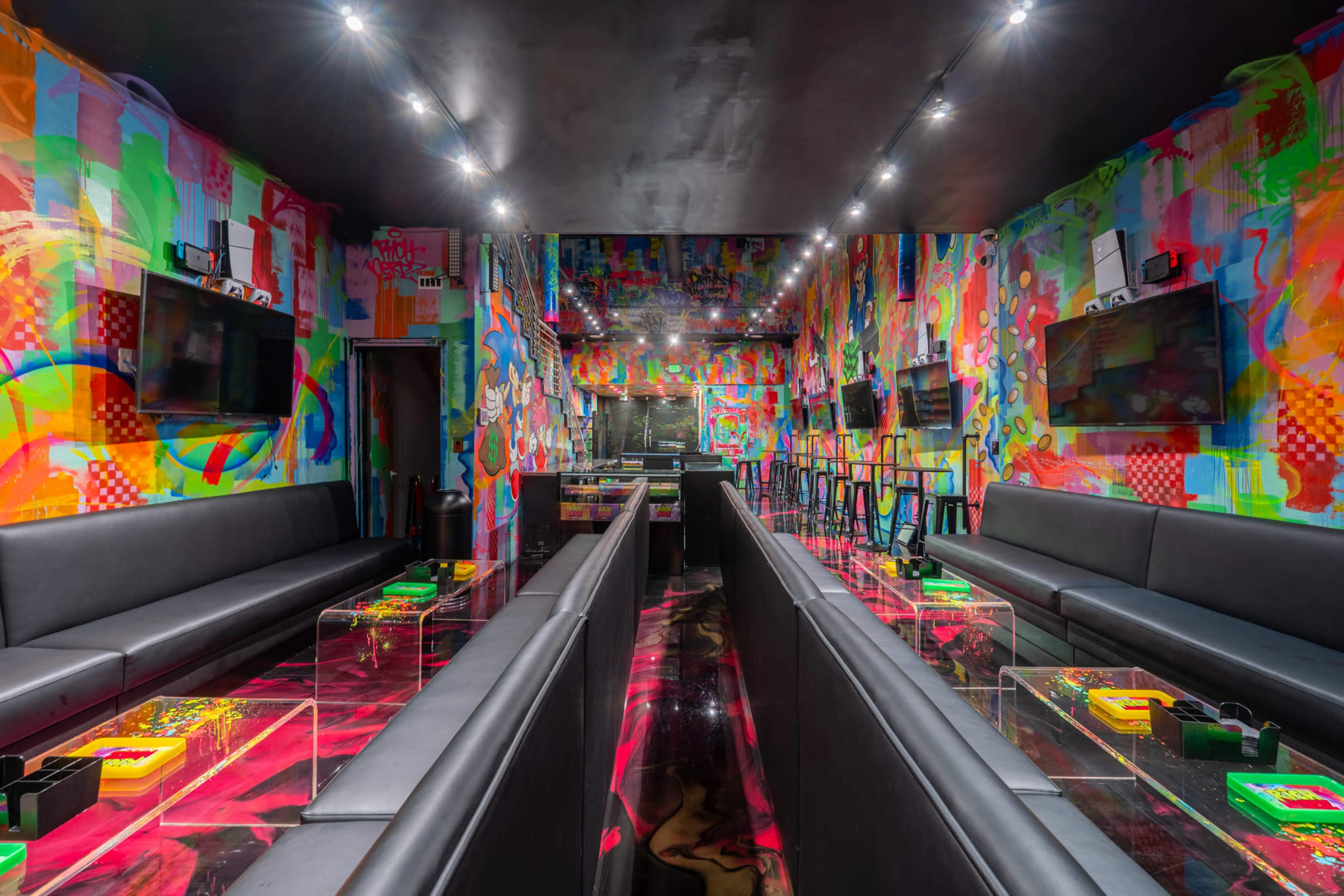 The interior of a vibrant gaming lounge features colorful graffiti-style walls, black seating along both sides, and illuminated tables.