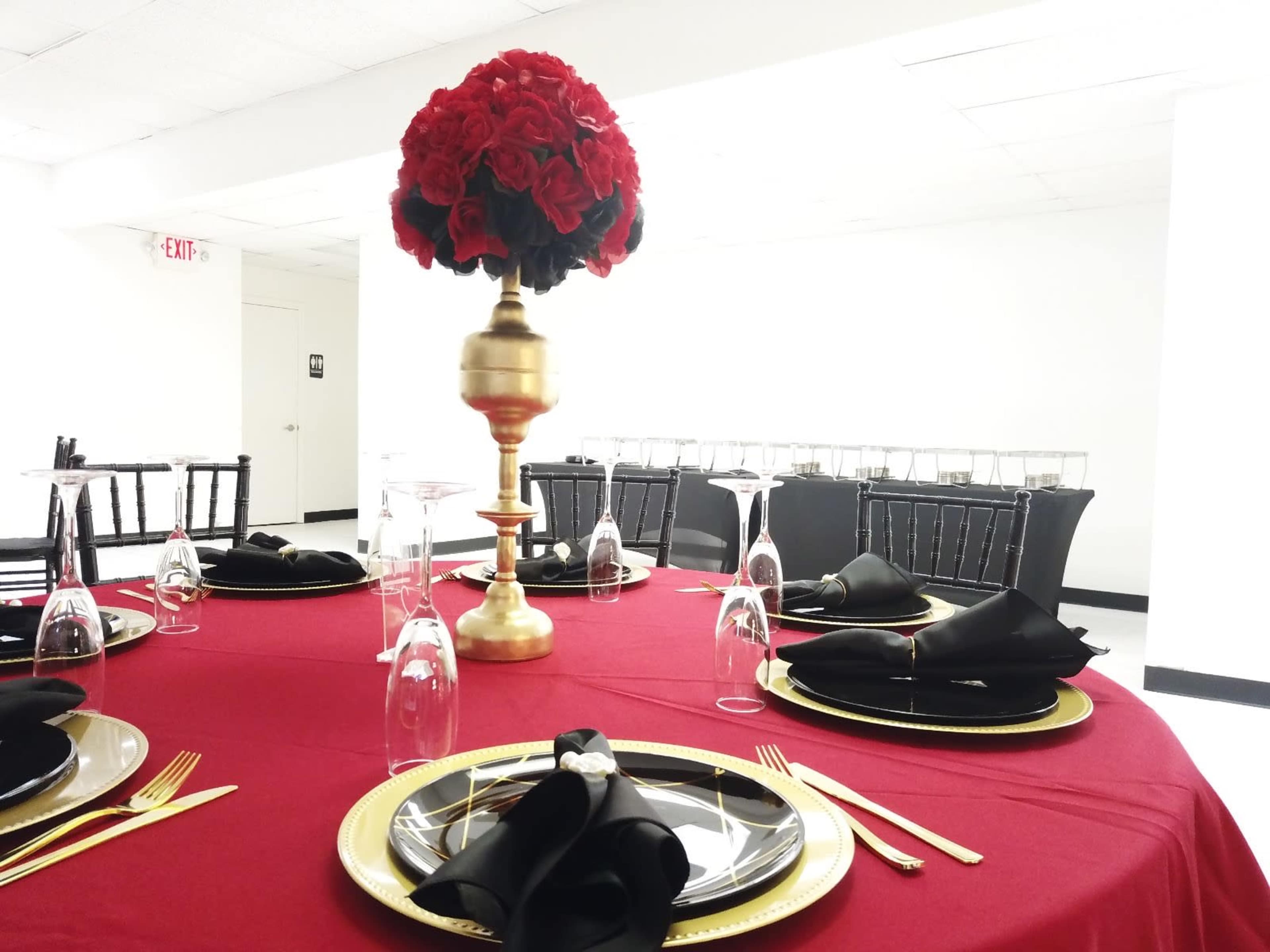 A round table is set with black and gold dinnerware, topped with a tall arrangement of red roses, against a white backdrop.
