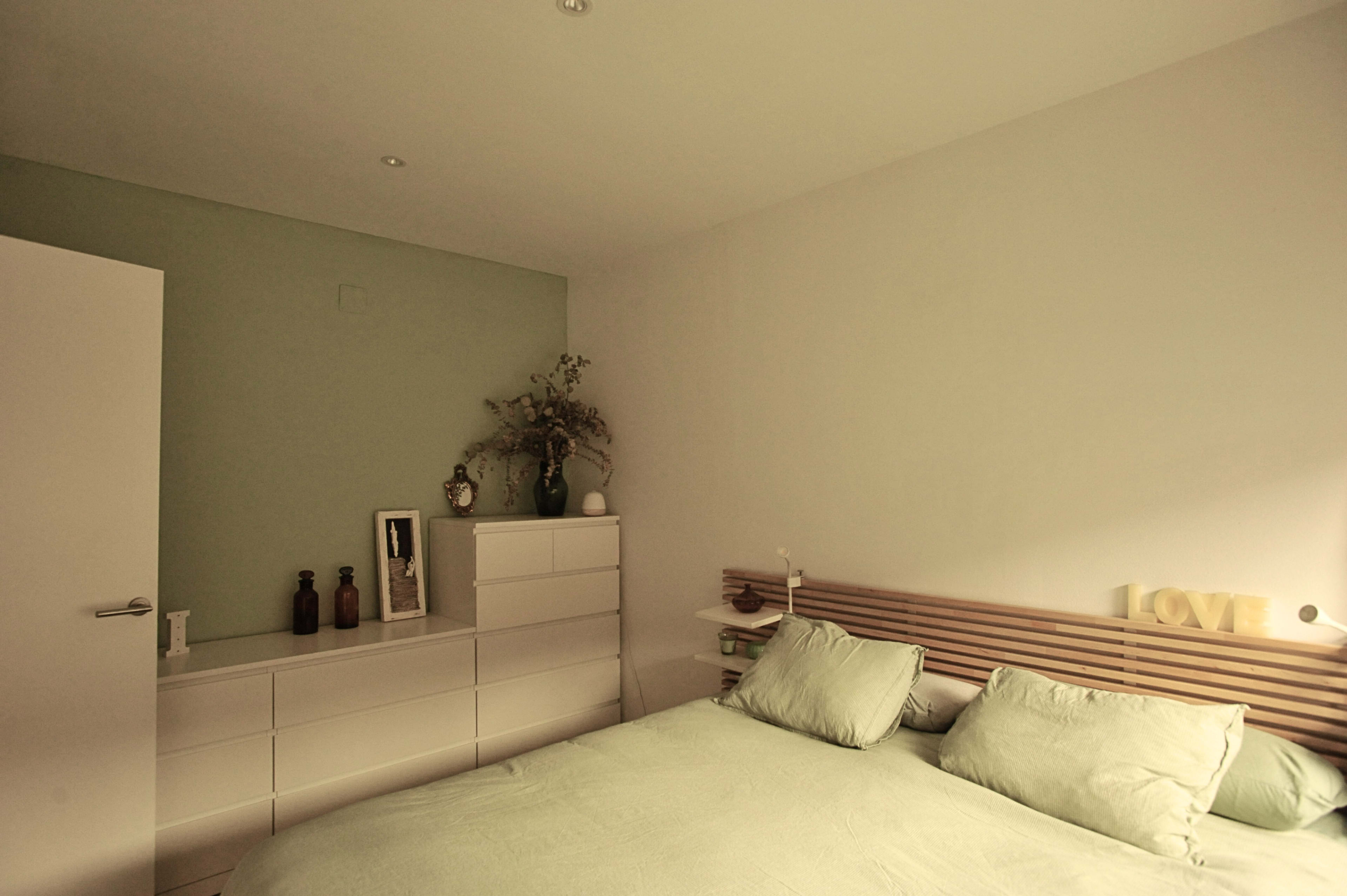 A minimalistic bedroom features a light green accent wall, a bed with soft linens, and a dresser adorned with decorative bottles and a mirror.