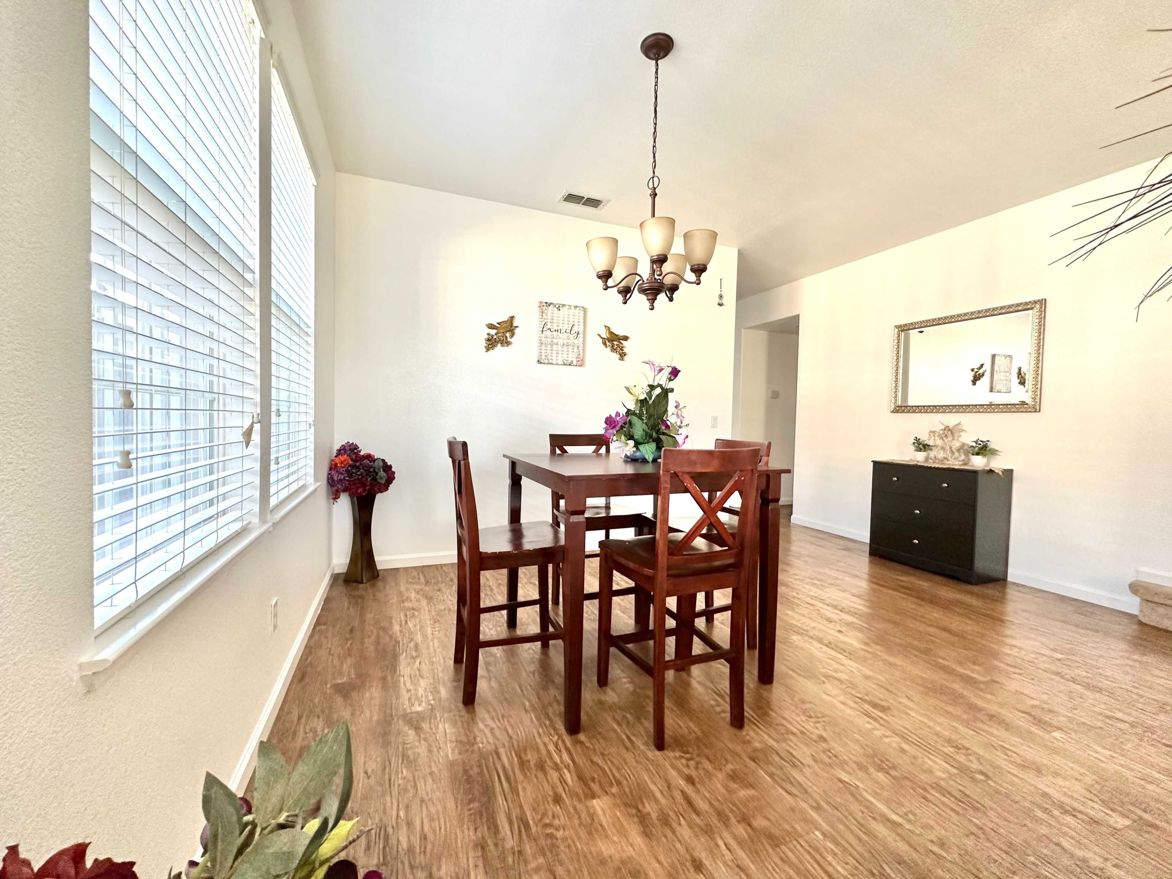 The image shows a dining area with a wooden table and four chairs, a chandelier overhead, and large windows allowing natural light, along with decorative elements on the walls and a sideboard nearby.