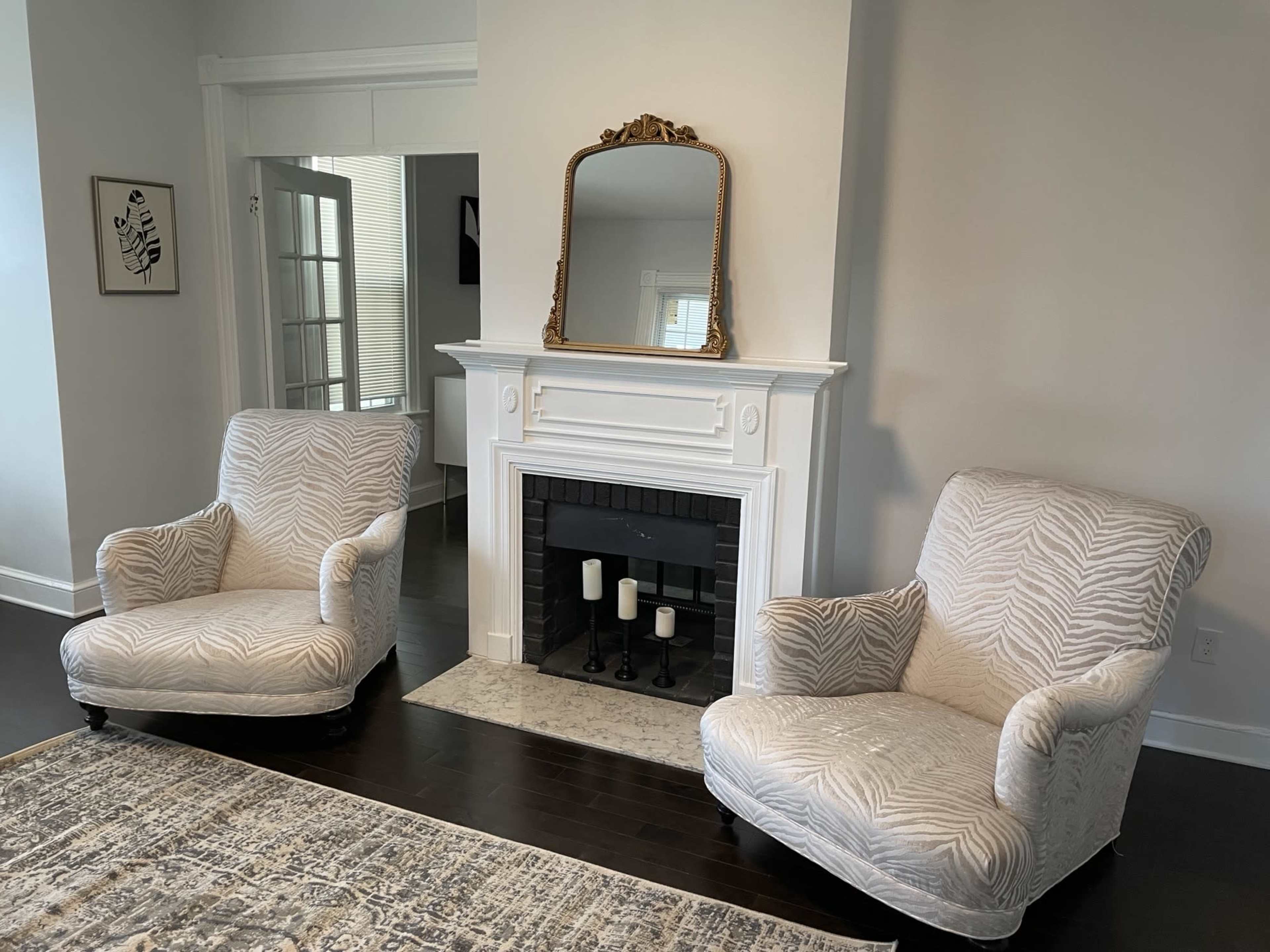 A living room with two patterned armchairs positioned near a white fireplace, which features a mirror above it and candles inside.
