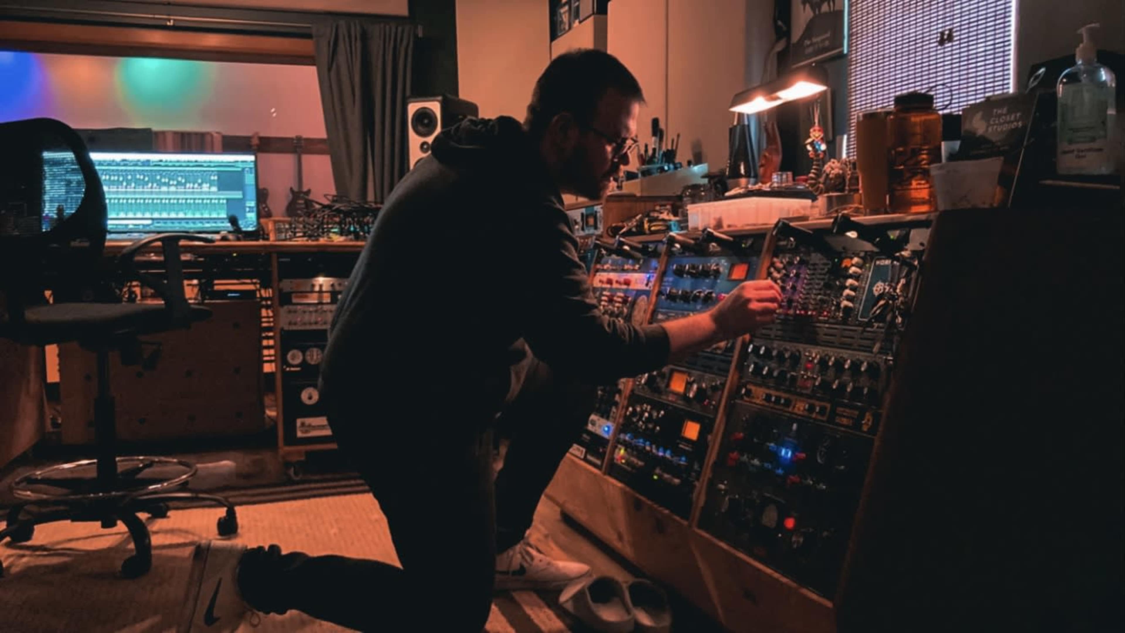 A man kneels in a dimly lit recording studio, adjusting knobs on a sound mixing console surrounded by various audio equipment.