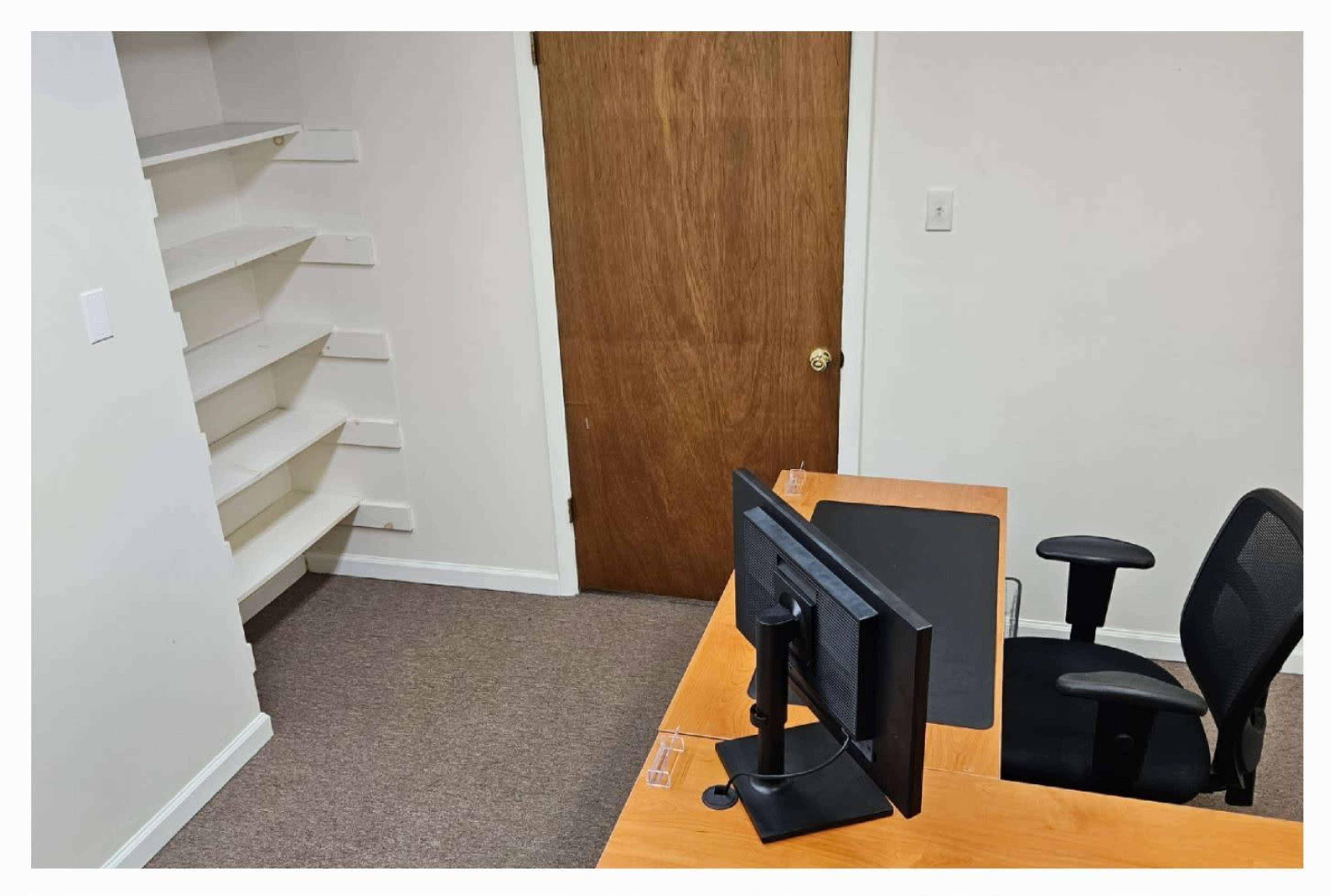 A small office space with a wooden desk, a computer monitor, a chair, and a built-in shelf against the wall.