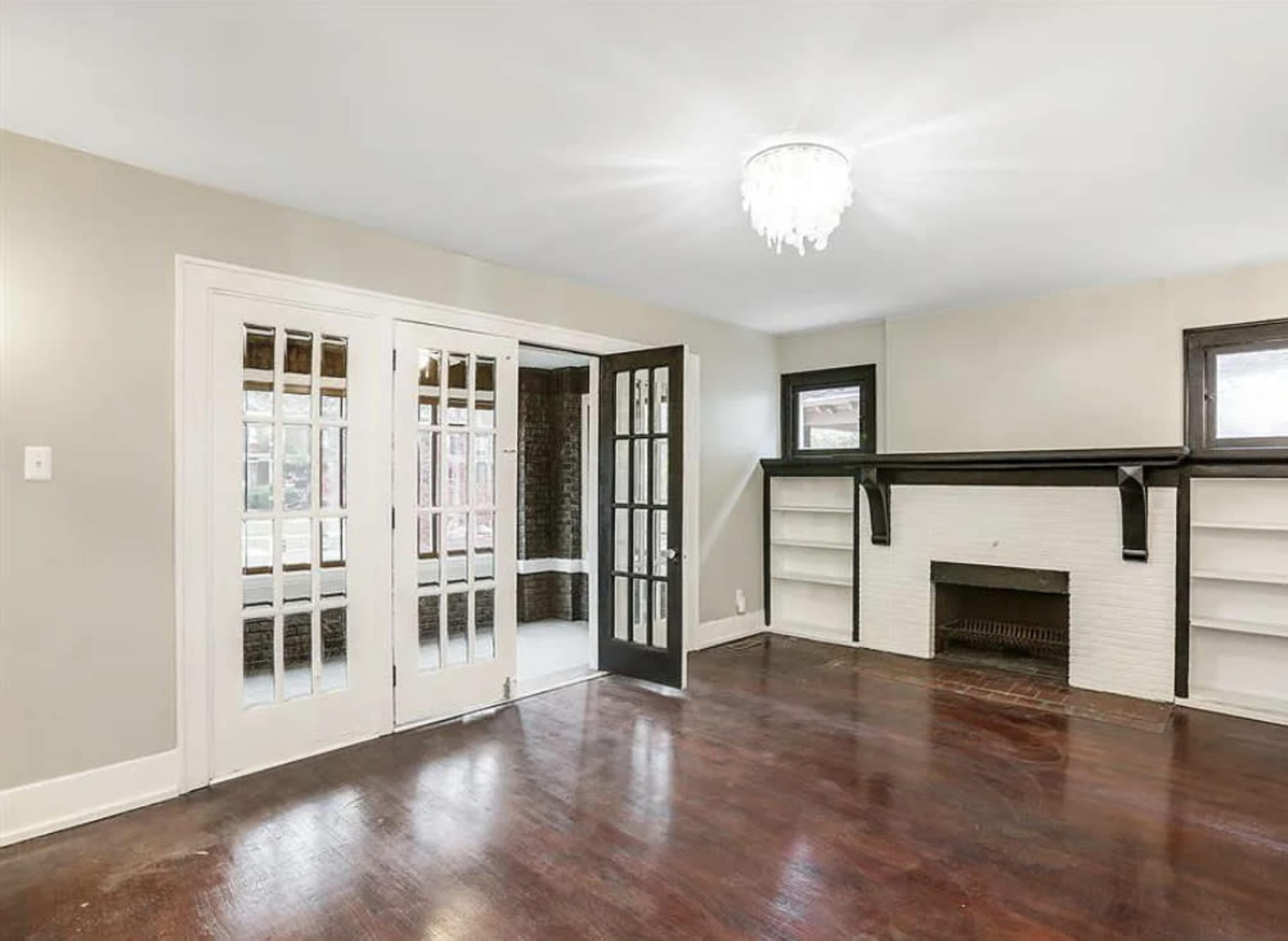 A spacious room with hardwood flooring, a white brick fireplace, and large double doors leading to another area.