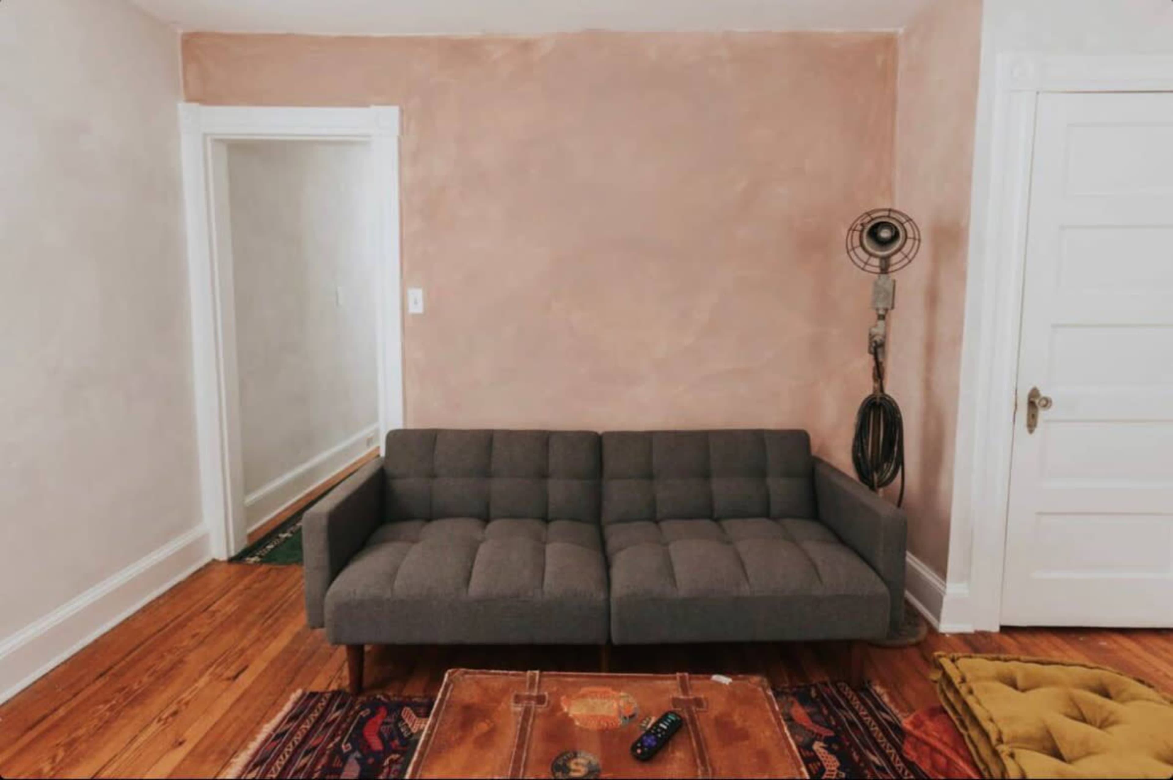 The image shows a modern gray sofa positioned in a room with light pink walls and wooden flooring, complemented by a vintage floor fan and a coffee table.