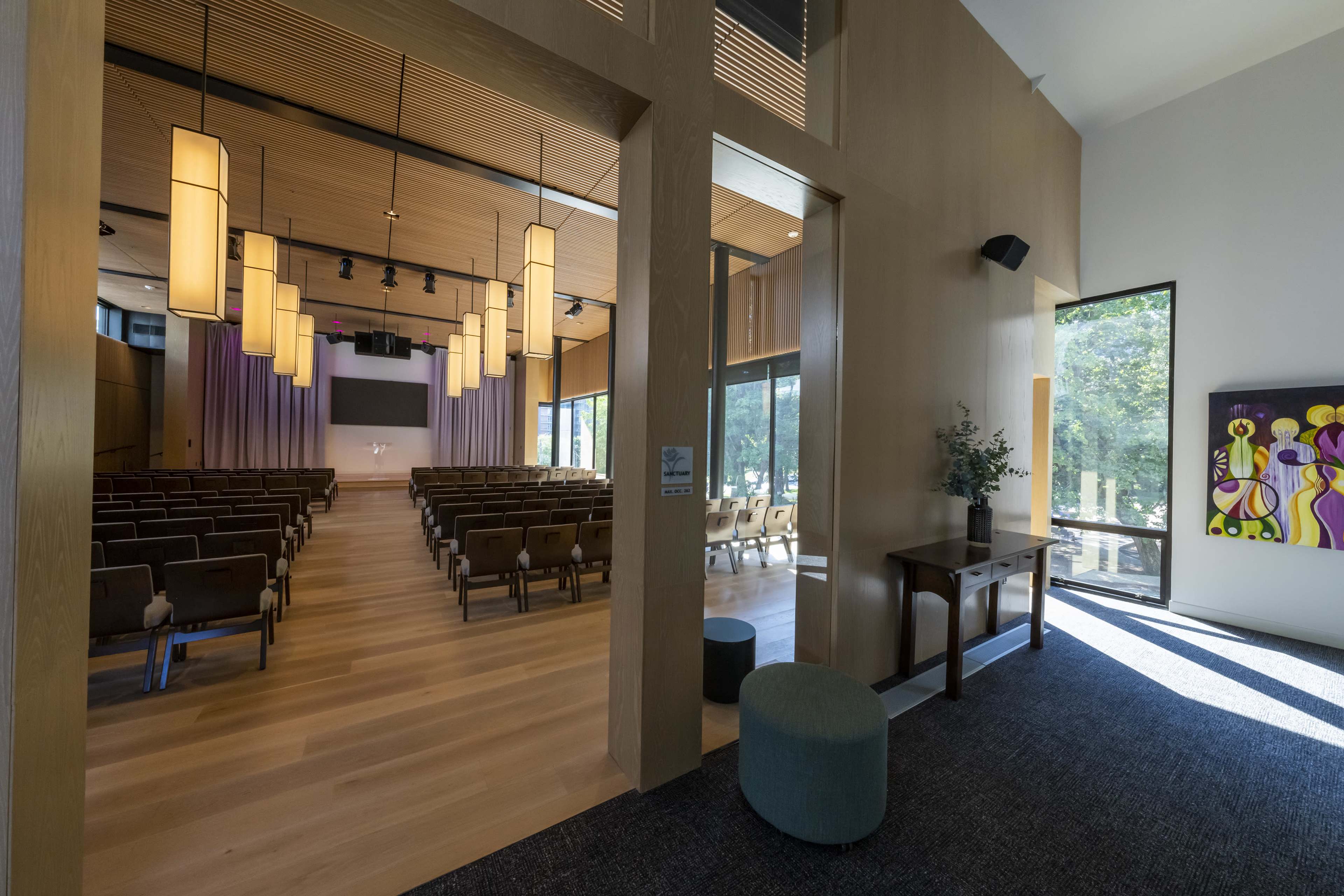 A modern auditorium with wooden walls and a stage, viewed from a hallway that features a console table and art on the wall.