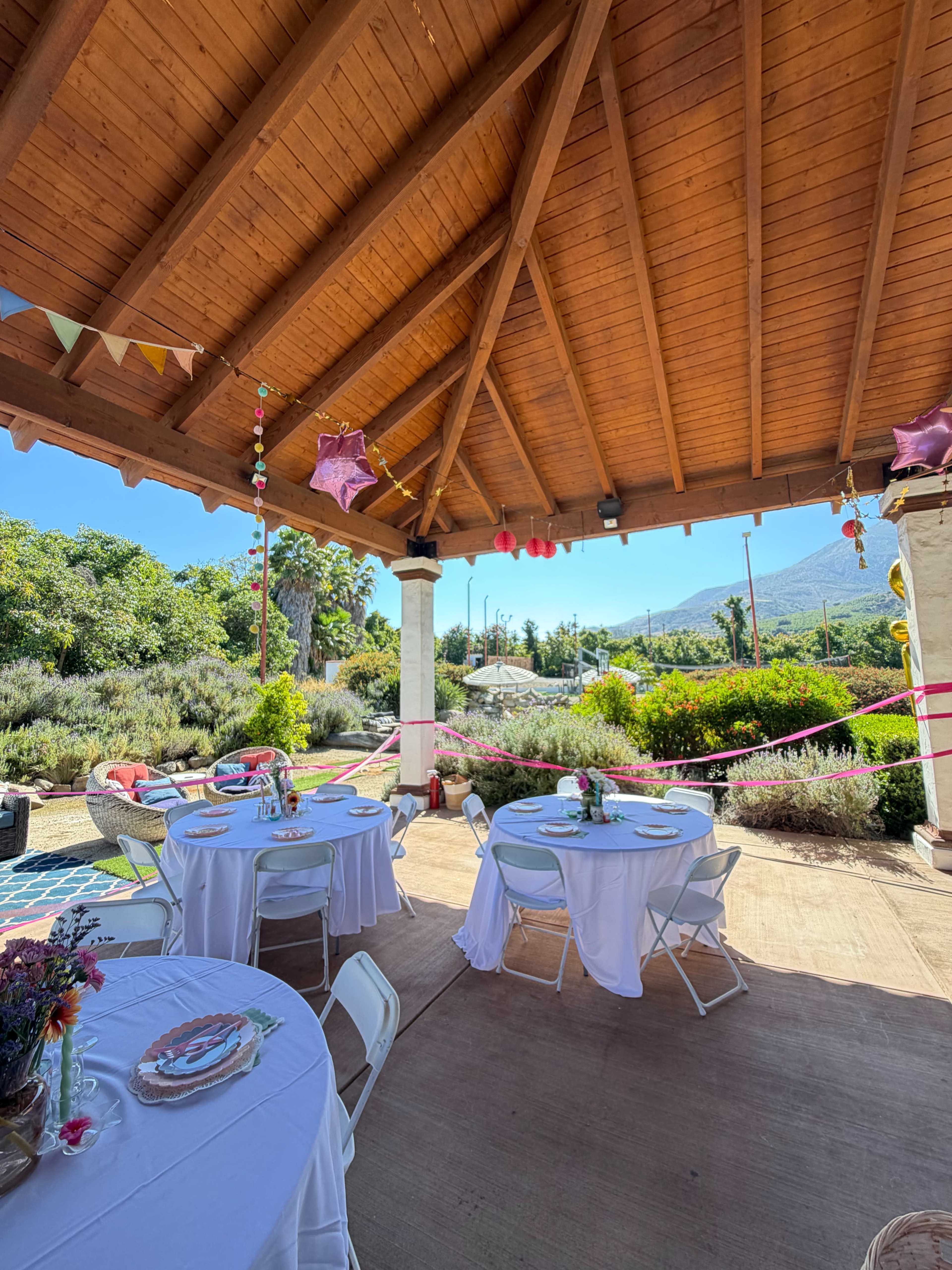 A covered outdoor setting features two round tables with white tablecloths, surrounded by greenery and decorated with streamers and balloons.