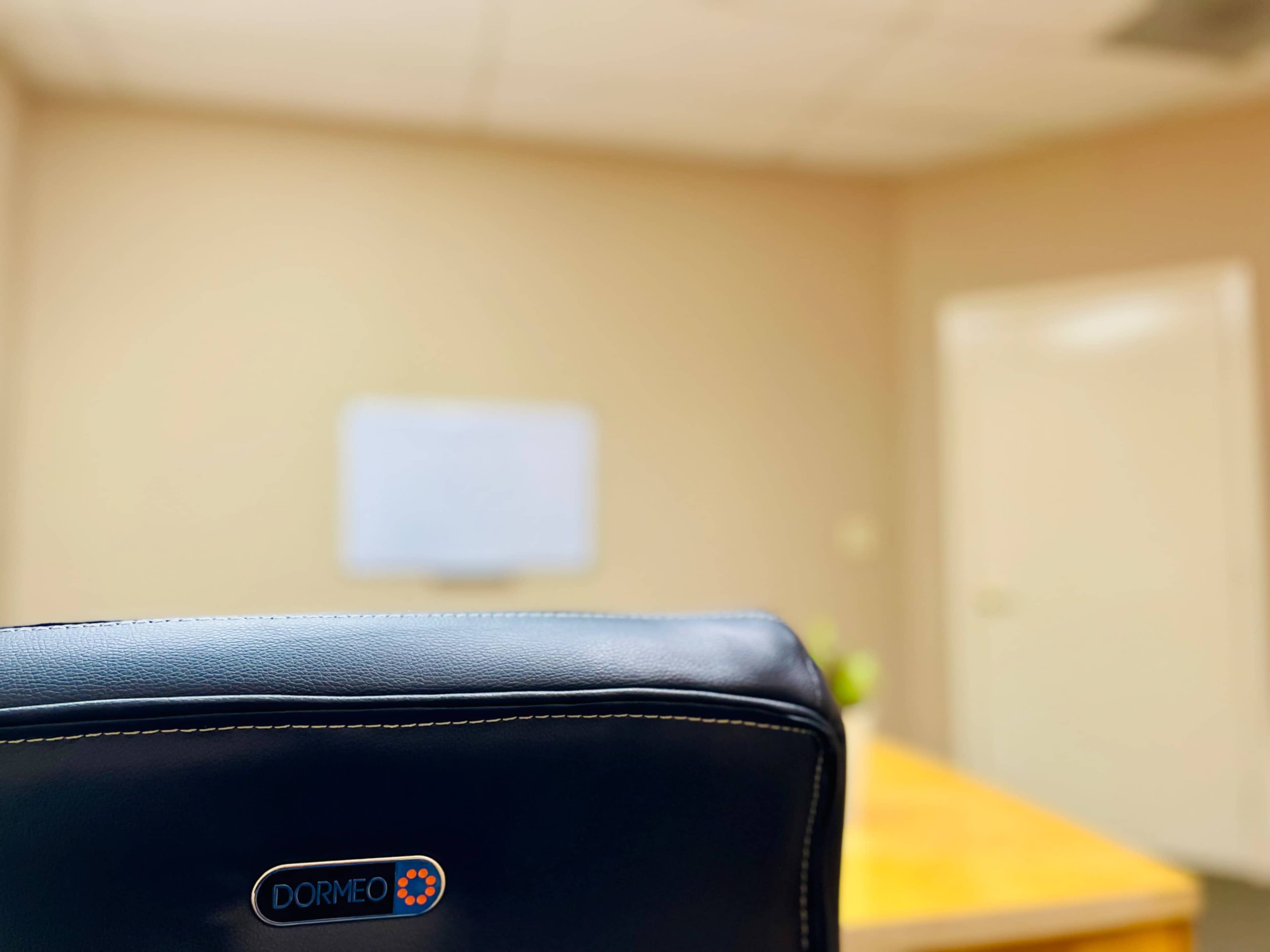 The image shows the back of an office chair labeled "DORMEO" facing a wooden table and a blank whiteboard in a softly lit office setting.