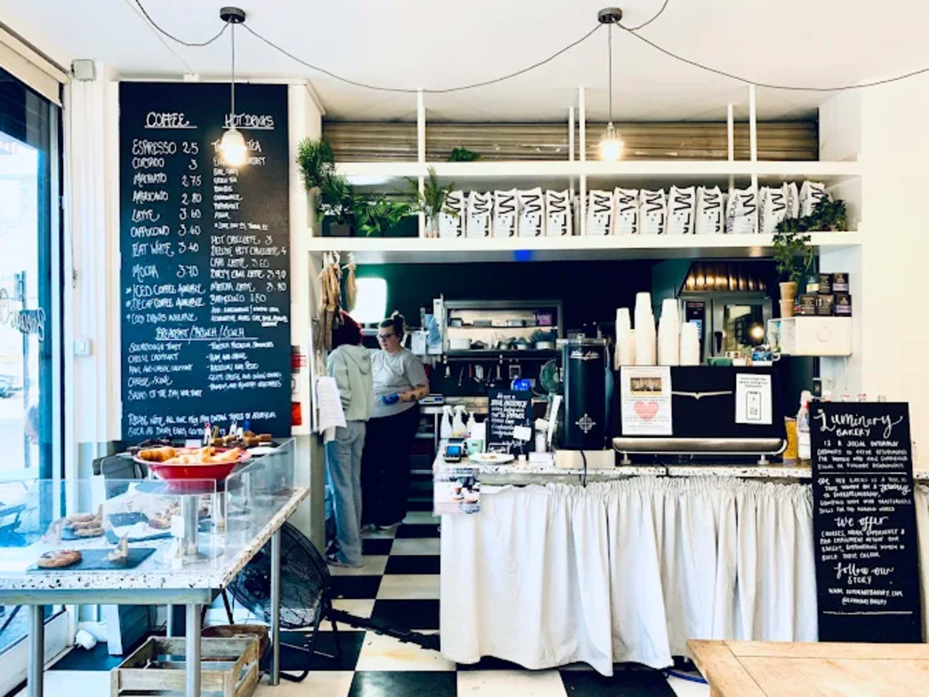 The image shows a cozy coffee shop interior with a chalkboard menu on the wall, a counter with coffee equipment, and pastries displayed in a glass case.