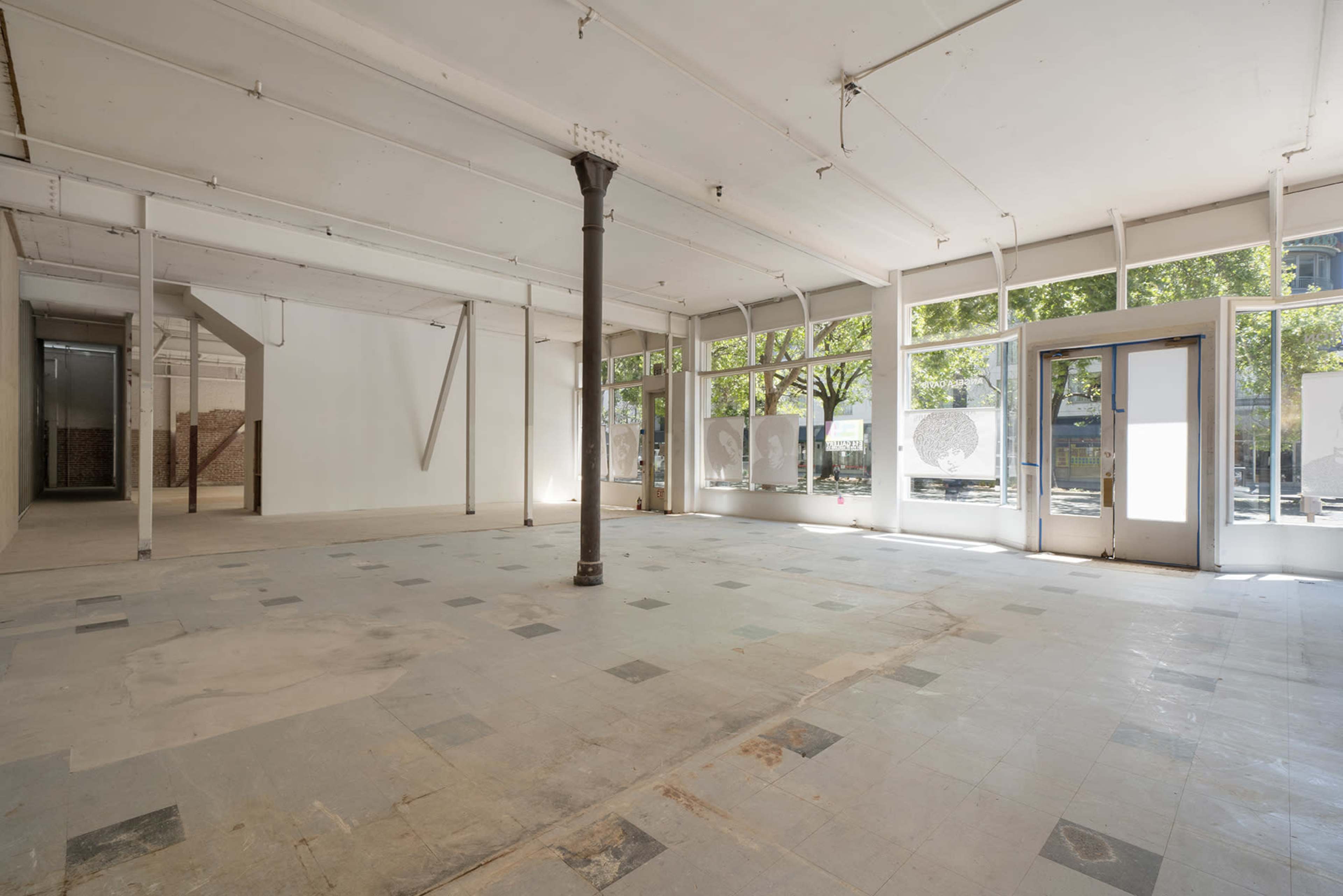 An empty commercial space with large windows and a central column, featuring tiled flooring and visible exterior views.