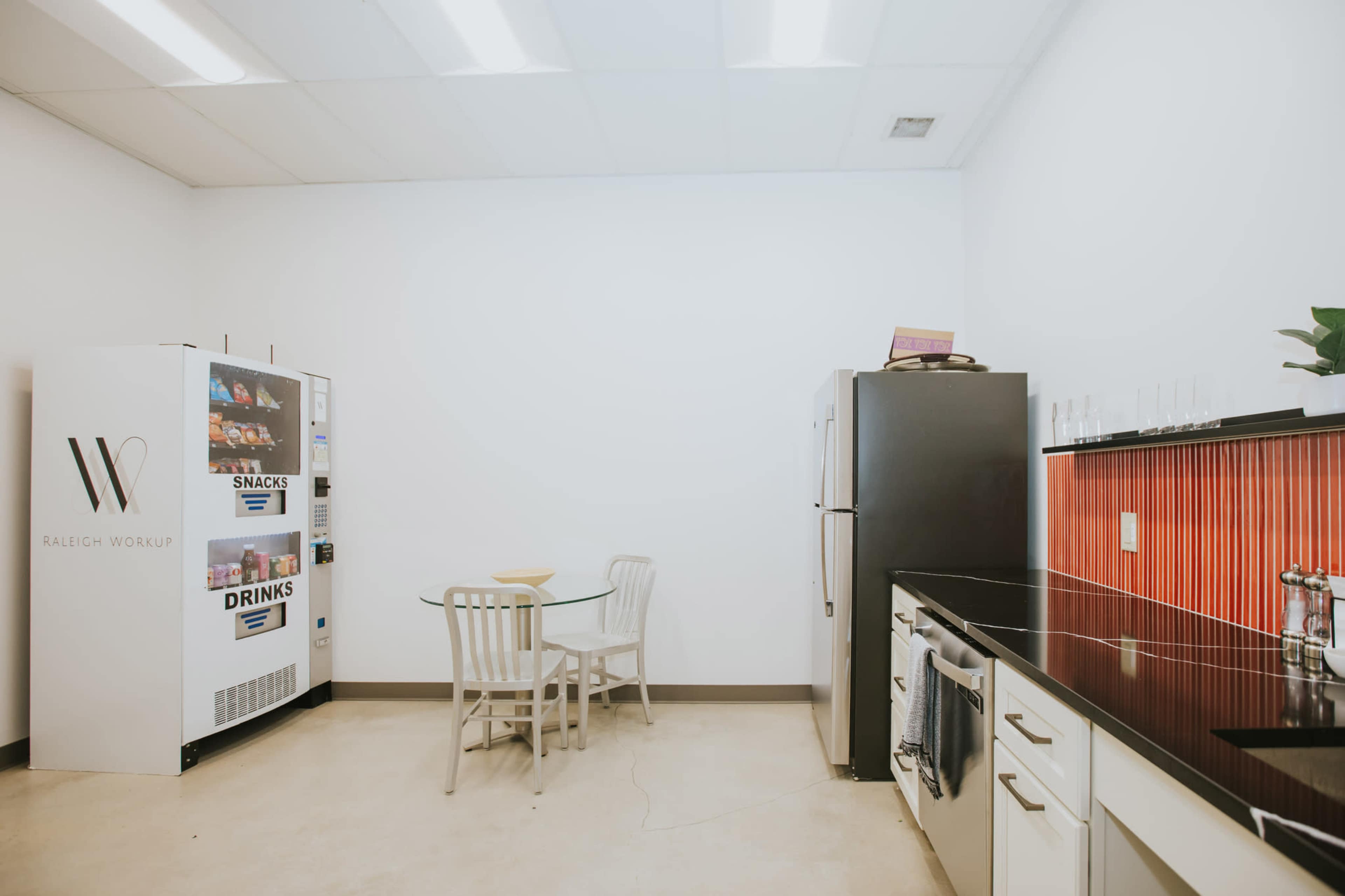 The image shows a small kitchen area with a table and chairs, a vending machine, and kitchen appliances against a white wall.