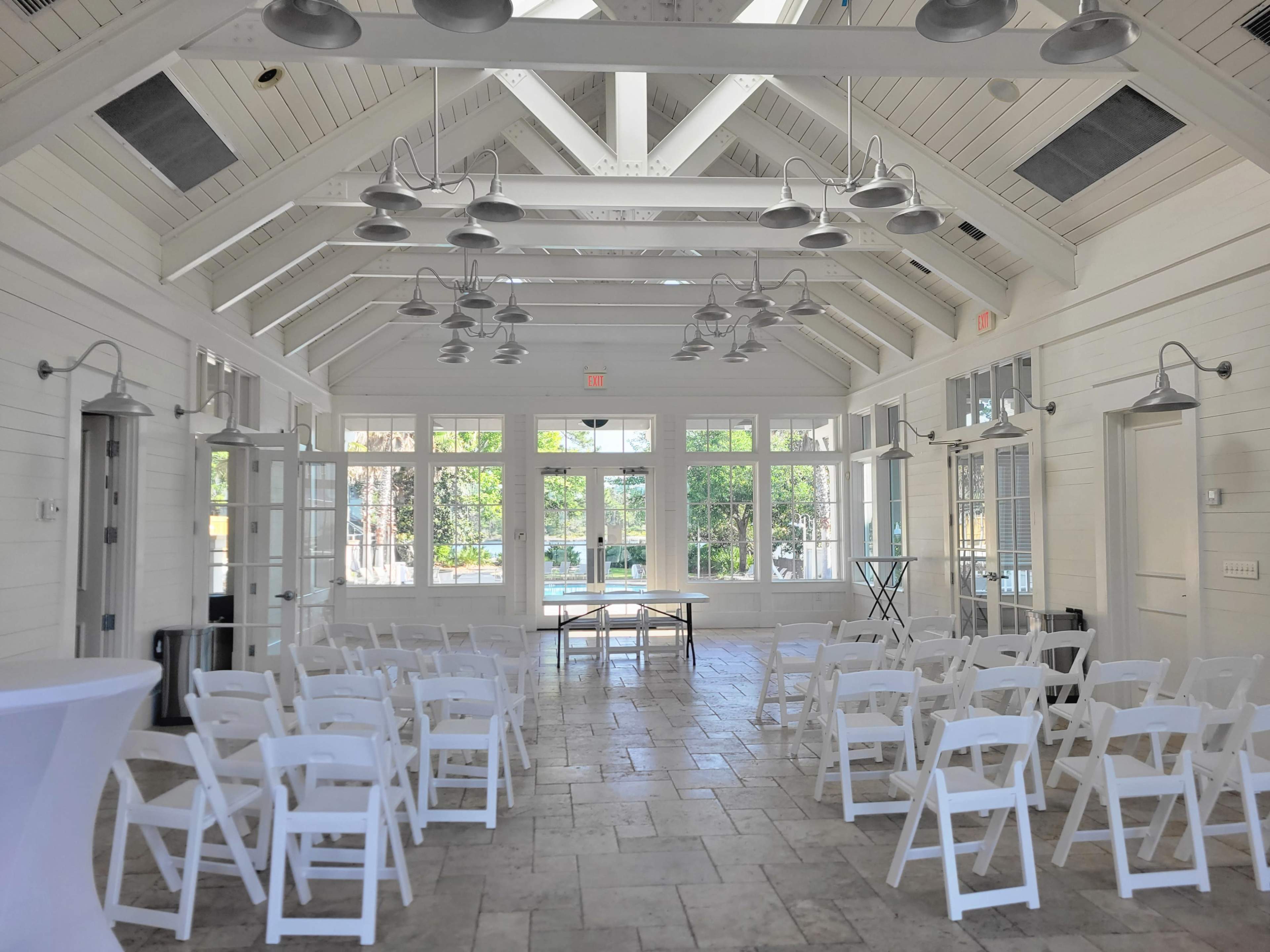 The image shows a bright, white-paneled event space with rows of white chairs set up facing a table and large windows.