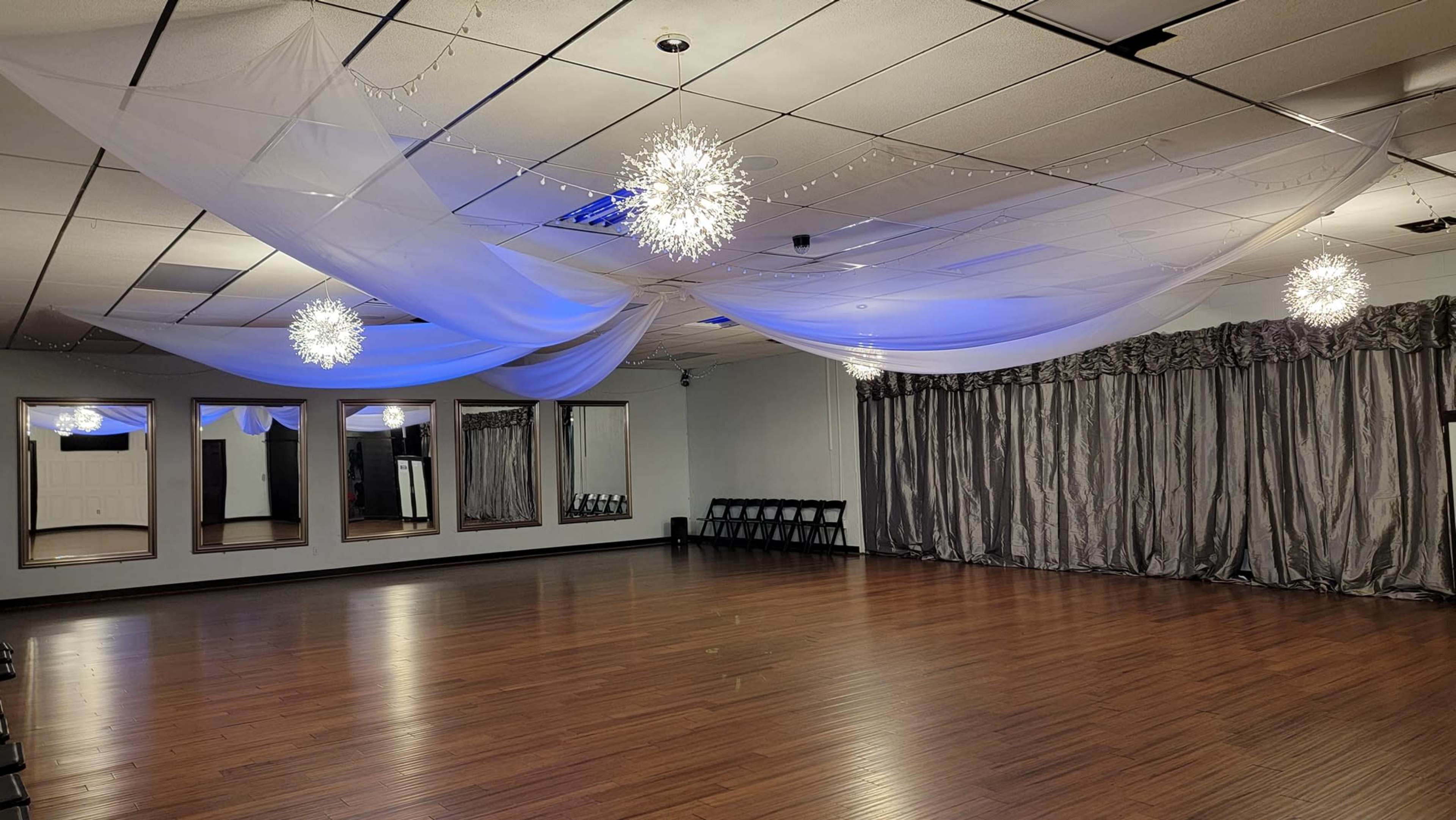 The interior of a dance studio features wooden floors, gray drapery, and decorative lighting installations hanging from the ceiling.