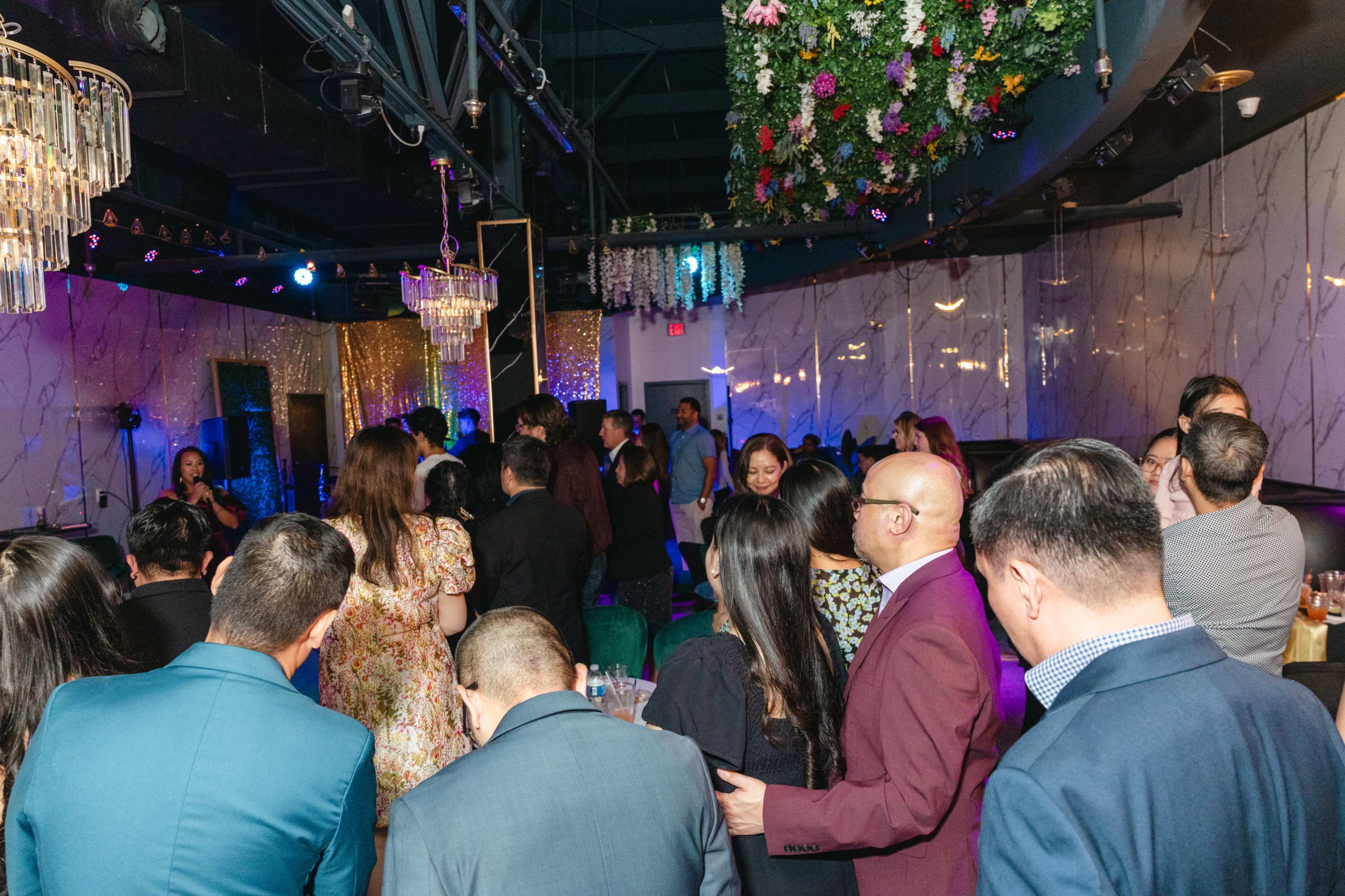 A crowd of people is gathered in a stylish, dimly lit venue with decorative flowers and chandeliers.