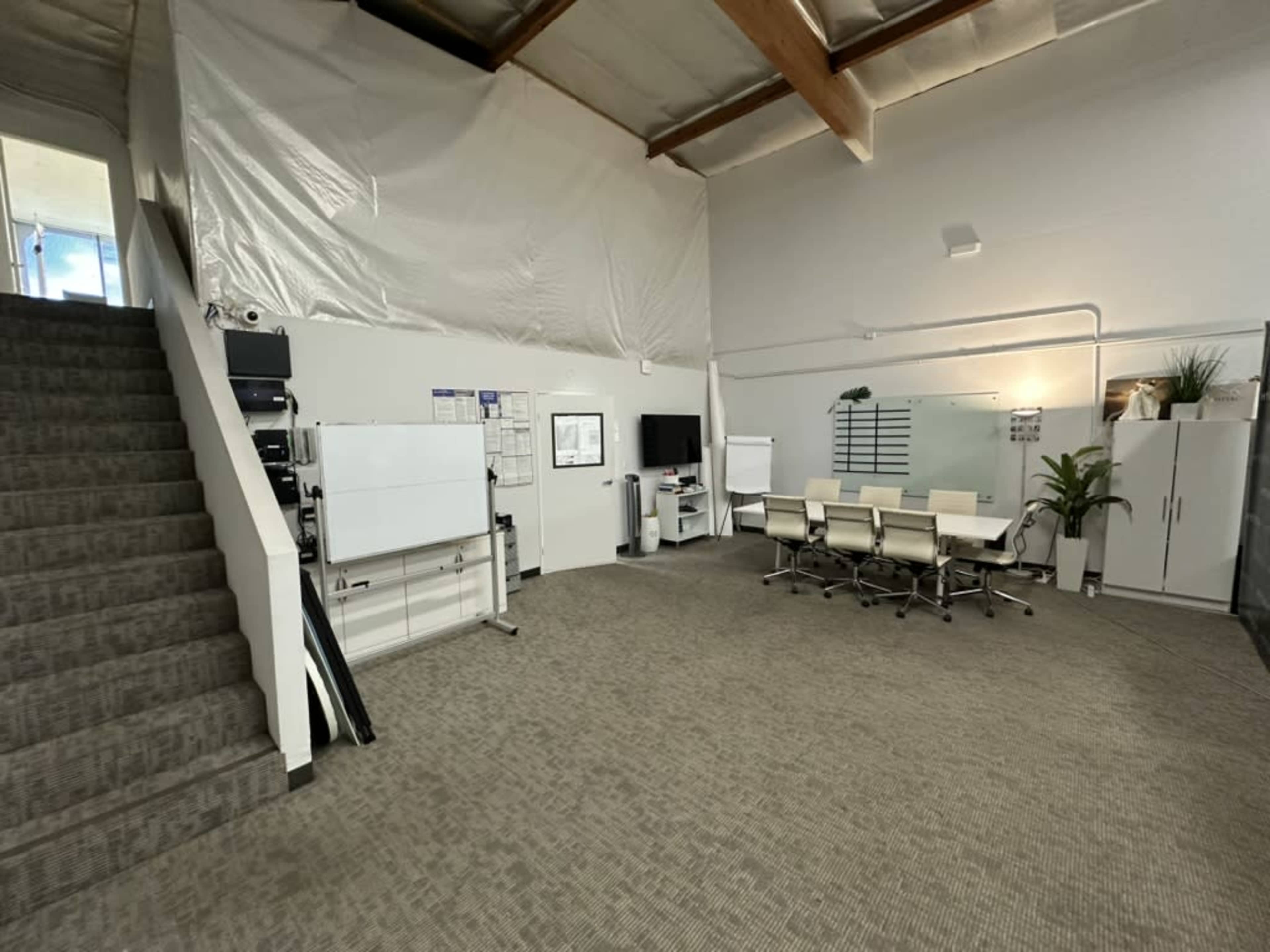 The image shows a modern office space featuring a staircase, a meeting area with a round table and chairs, a whiteboard, and several storage units.