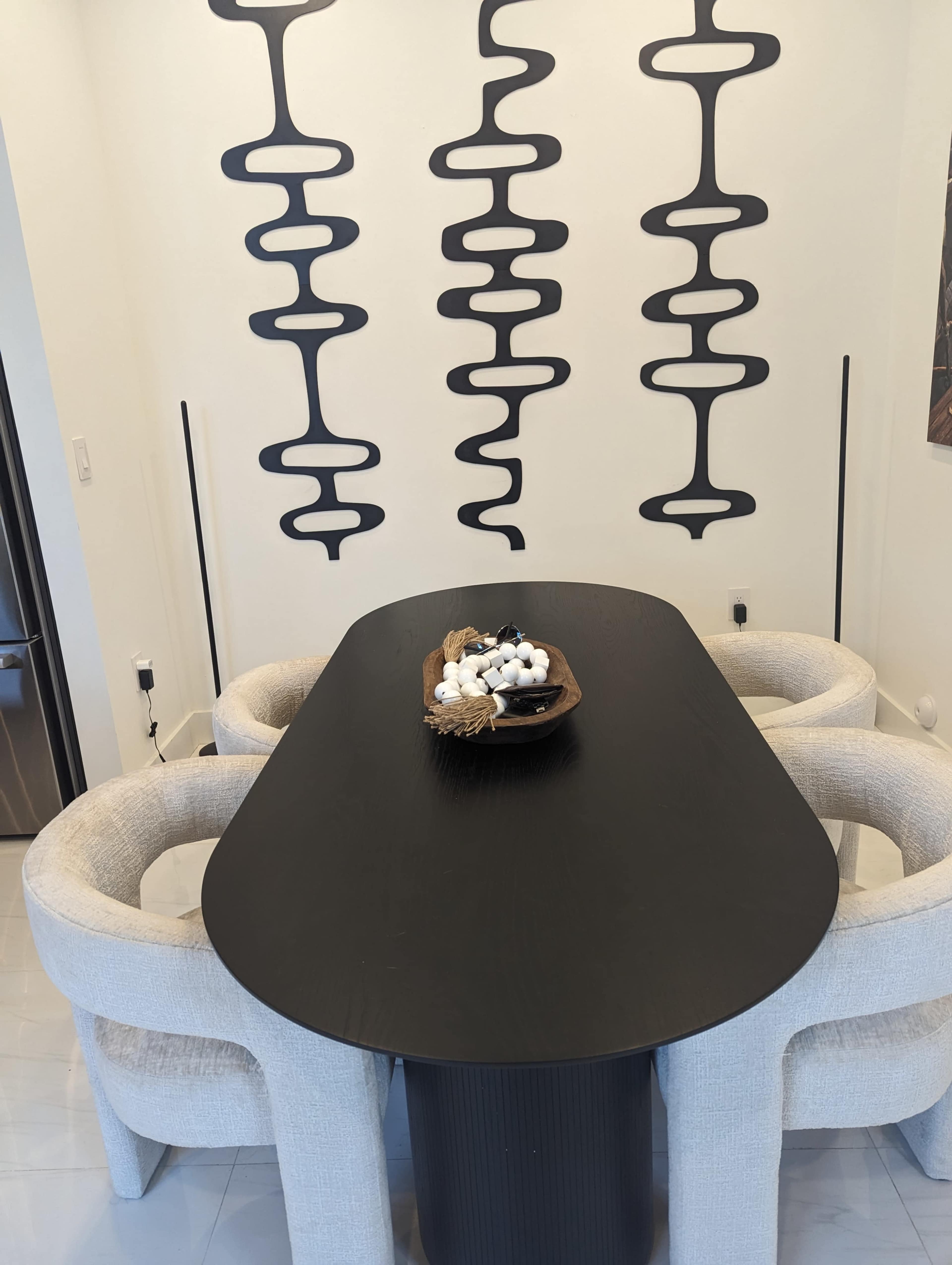 A modern dining area features a black oval table surrounded by two beige upholstered chairs, with abstract black designs on the wall behind.