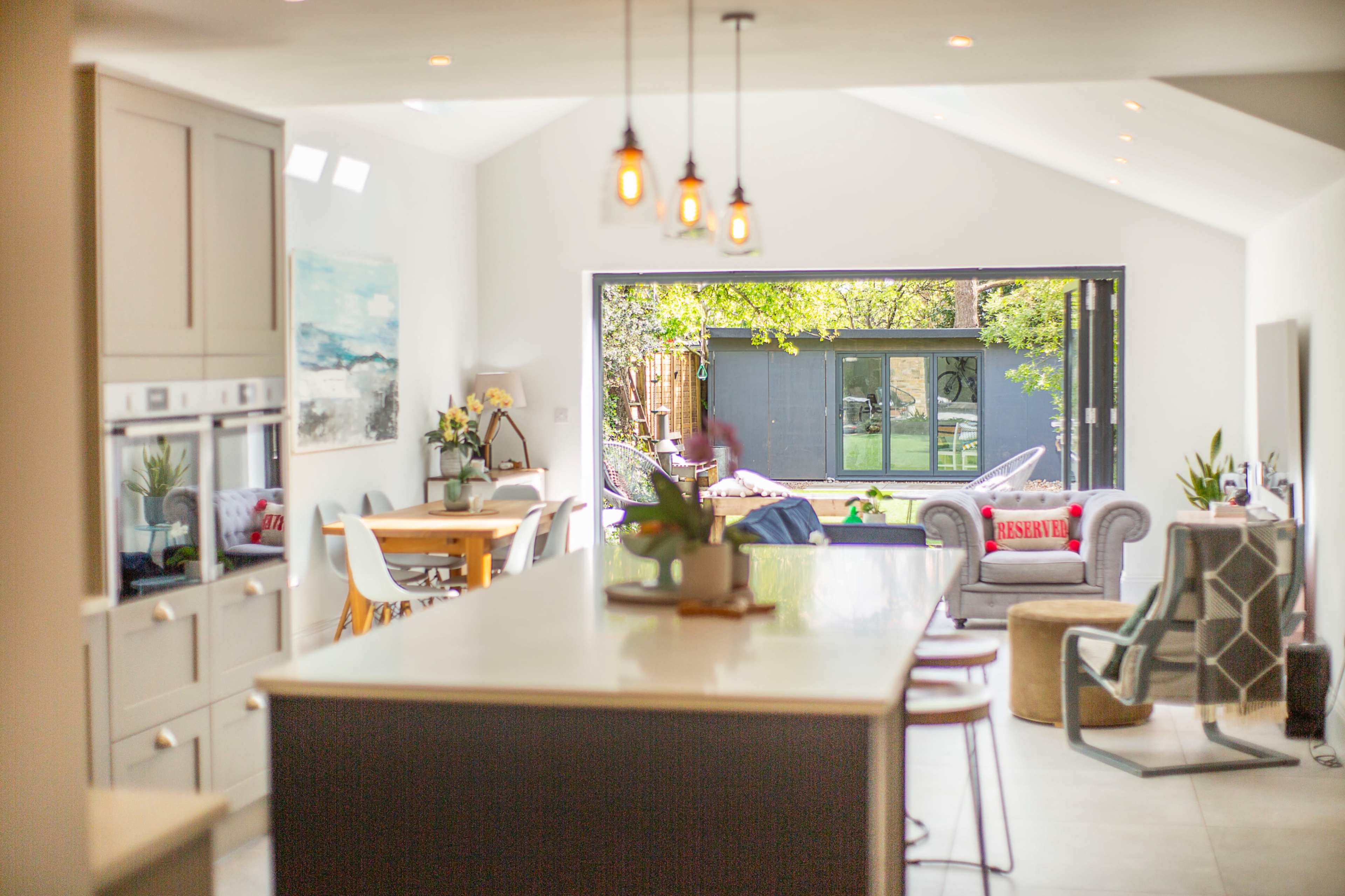The image shows a modern kitchen with a central island, leading to a spacious outdoor area through large glass doors.