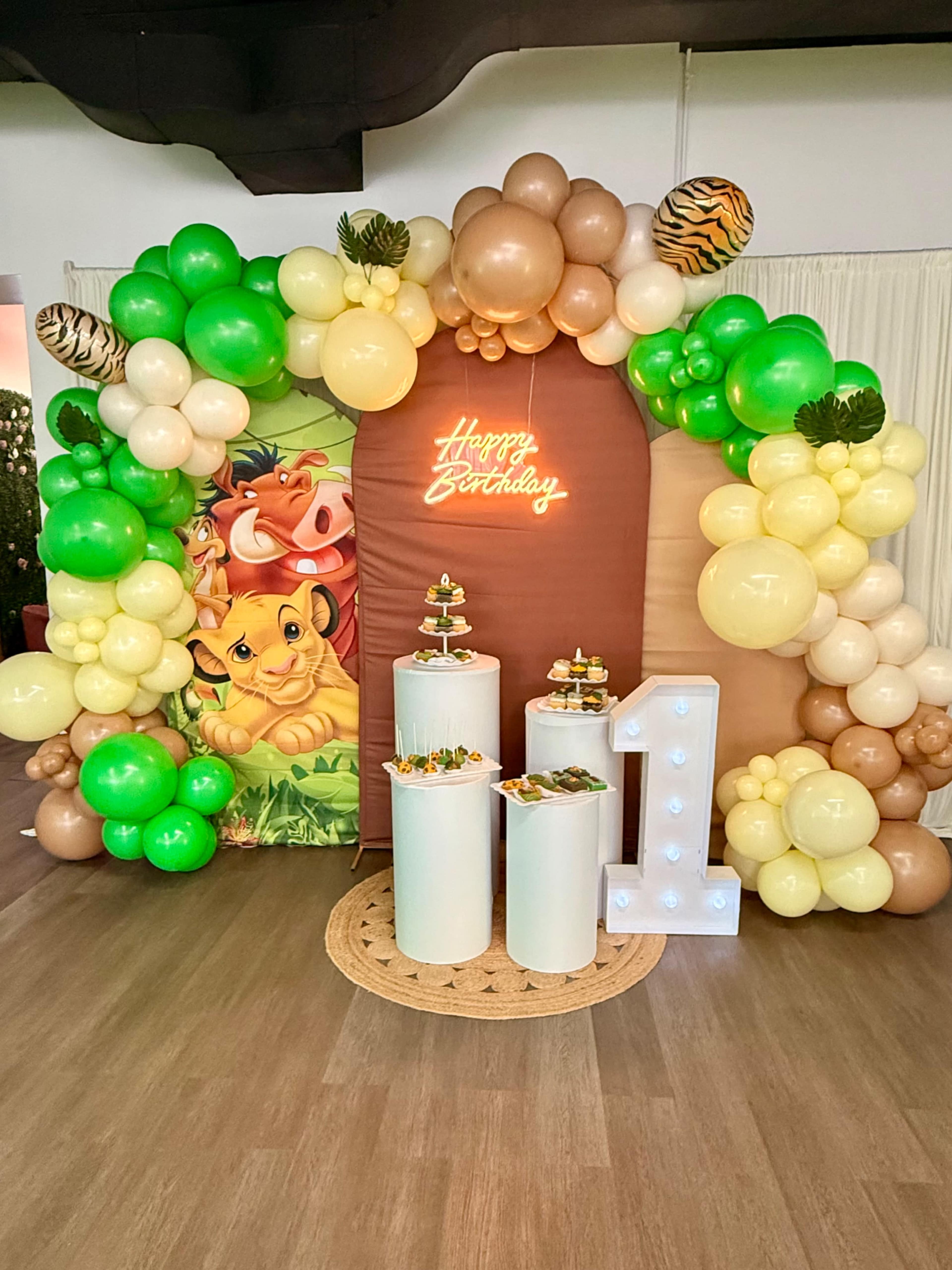 The scene features a festive birthday backdrop decorated with balloons in shades of green, beige, and gold, along with a neon "Happy Birthday" sign and a large white numeral "1" on display.