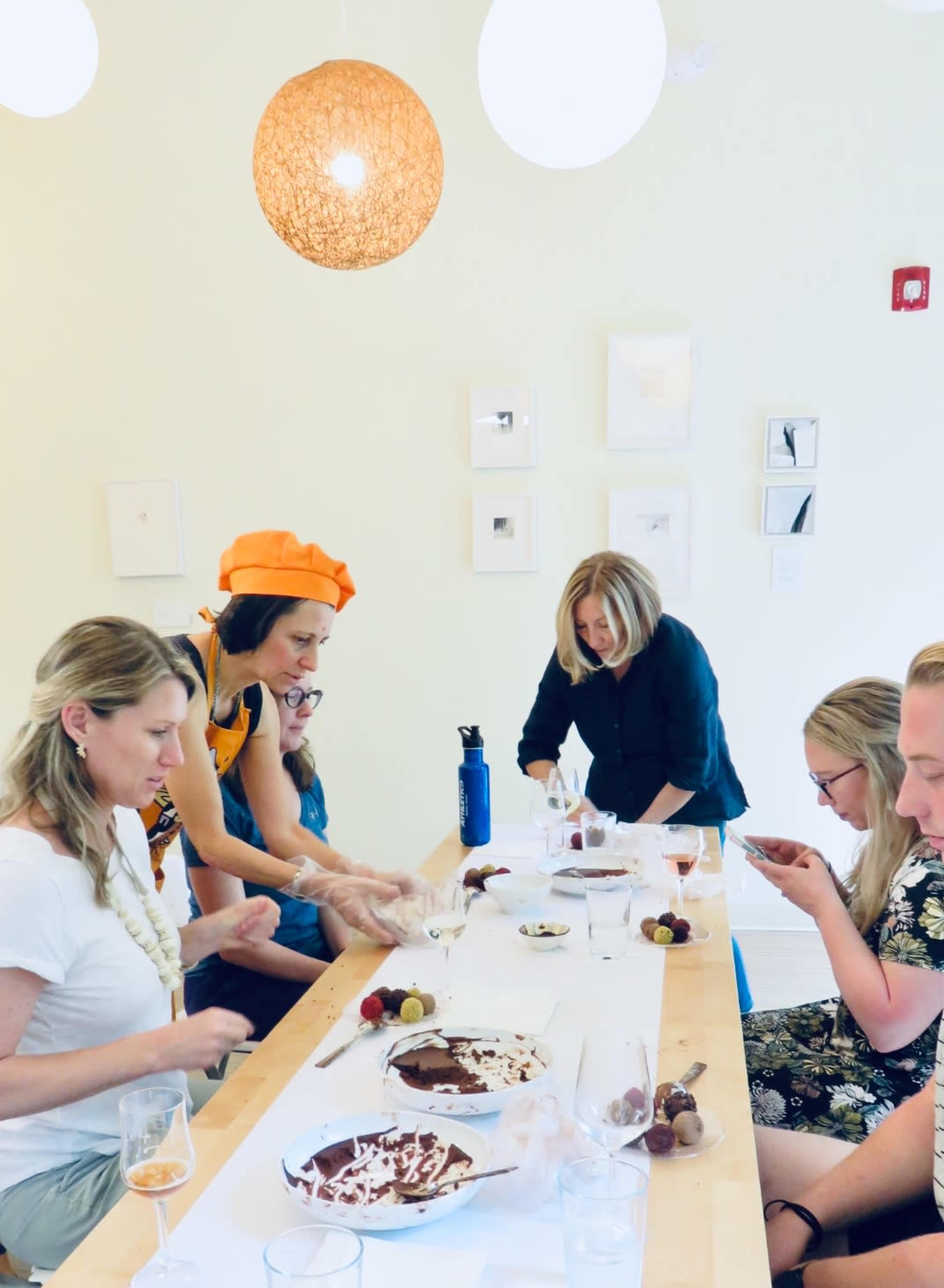 A group of six people are engaged in a chocolate-making class at a long table, with assorted chocolate desserts and a variety of tools and ingredients laid out.