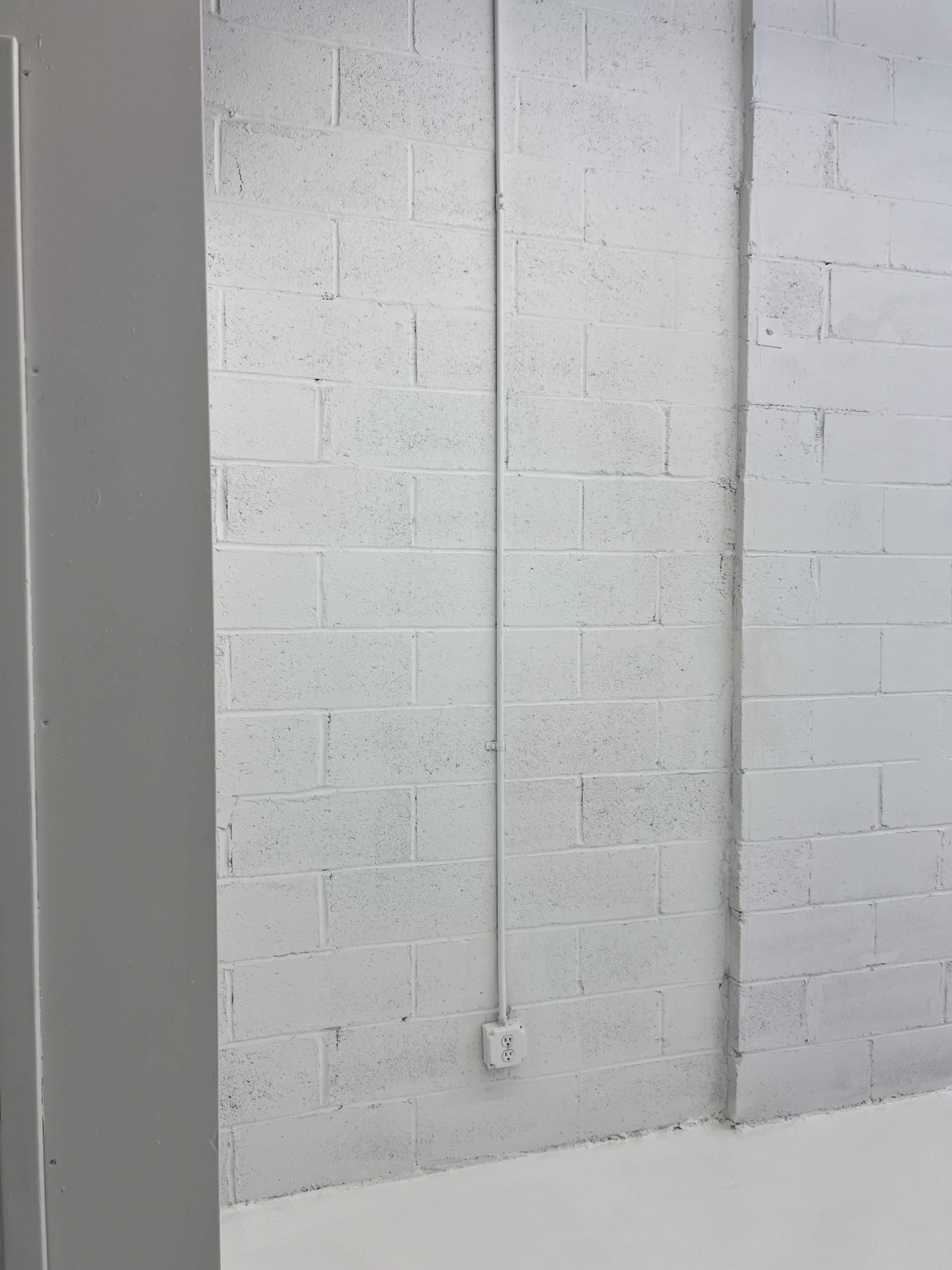 A blank, gray wall made of cinder blocks with a vertical electrical conduit and an outlet on the lower part.
