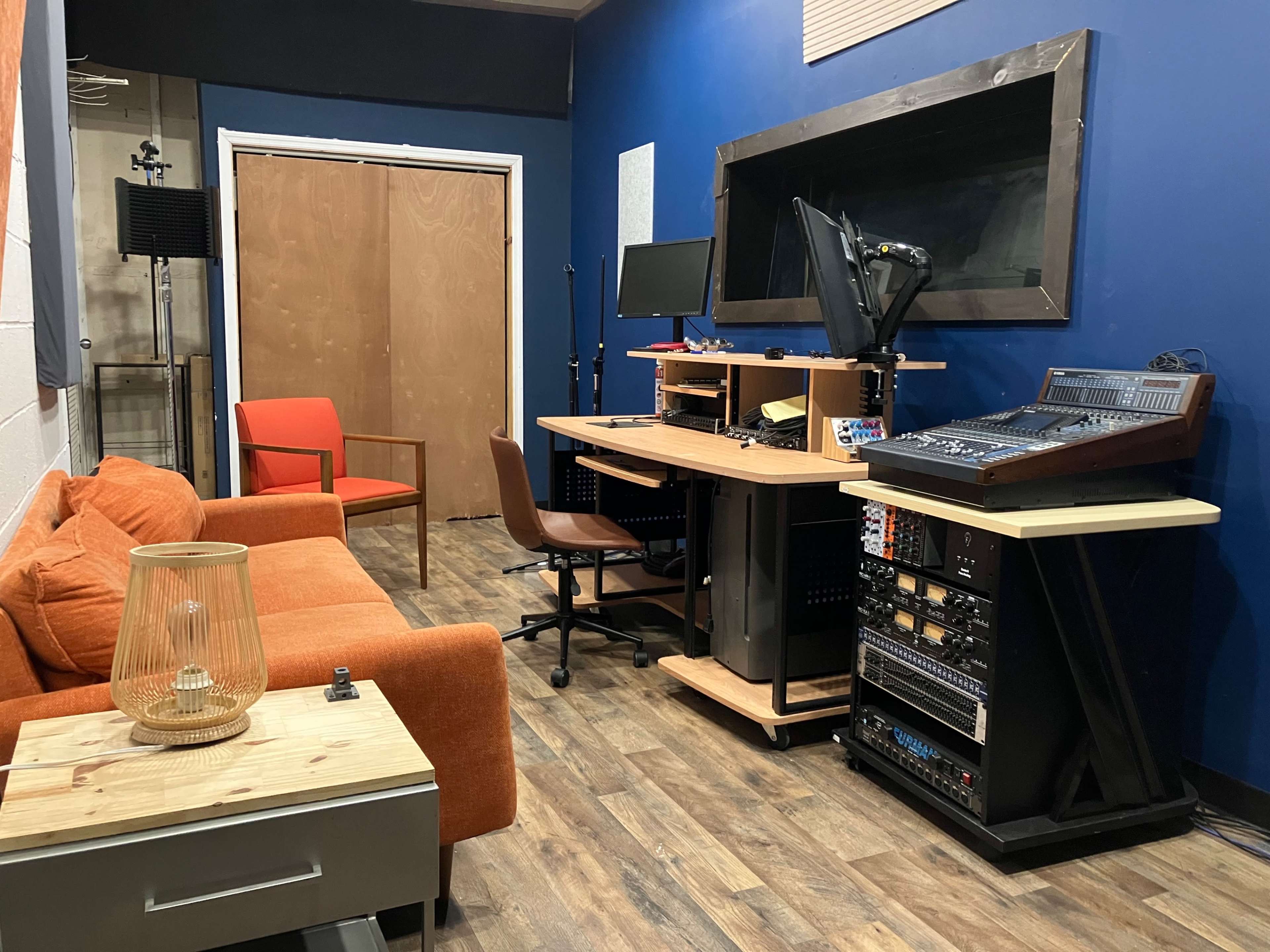 The image shows a small recording studio with an orange couch, a wooden desk with audio equipment, a chair, and a large window in a blue-walled room.