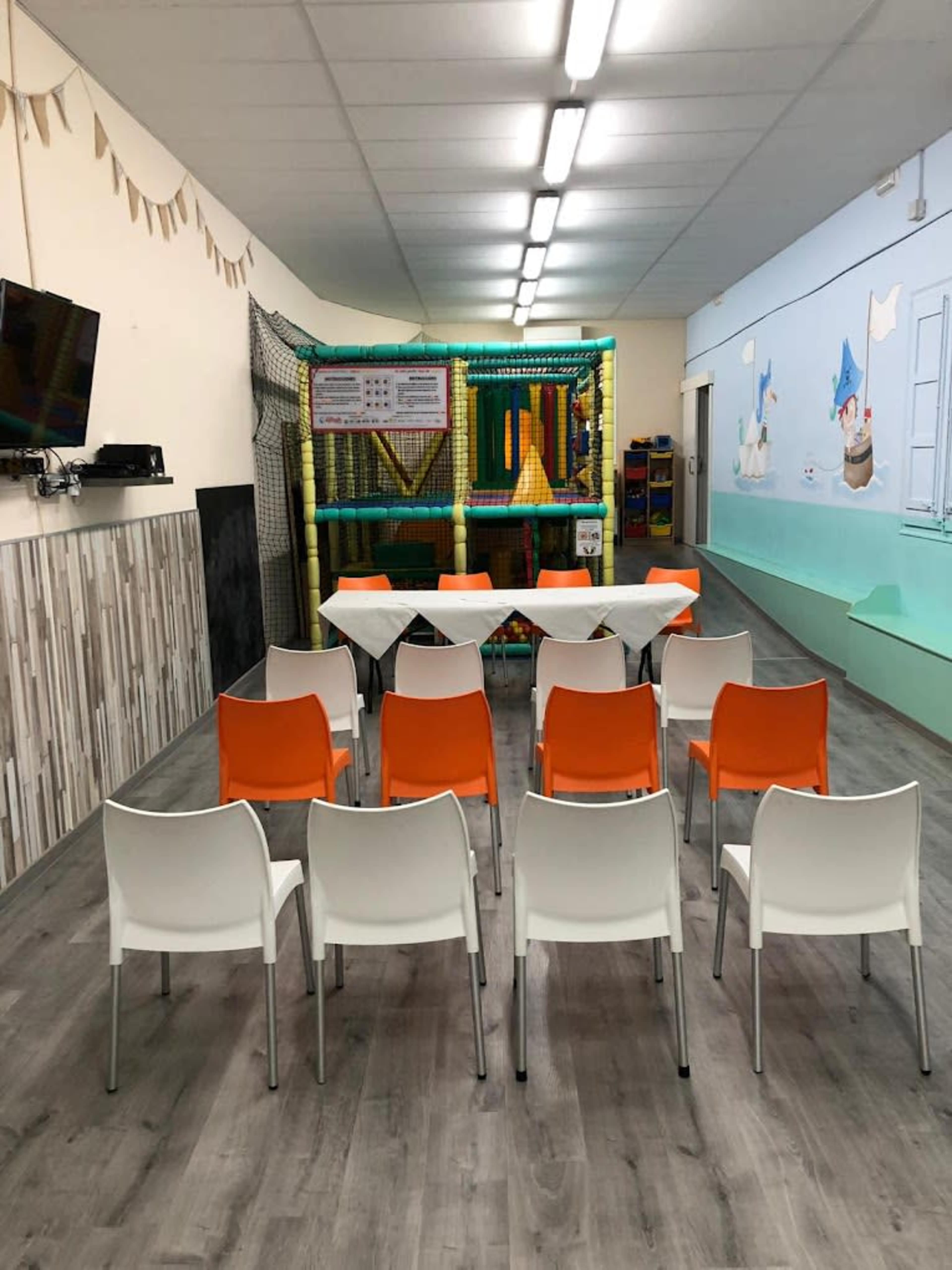 The image shows a spacious room with a play area in the back, arranged with a row of white and orange chairs facing a table.