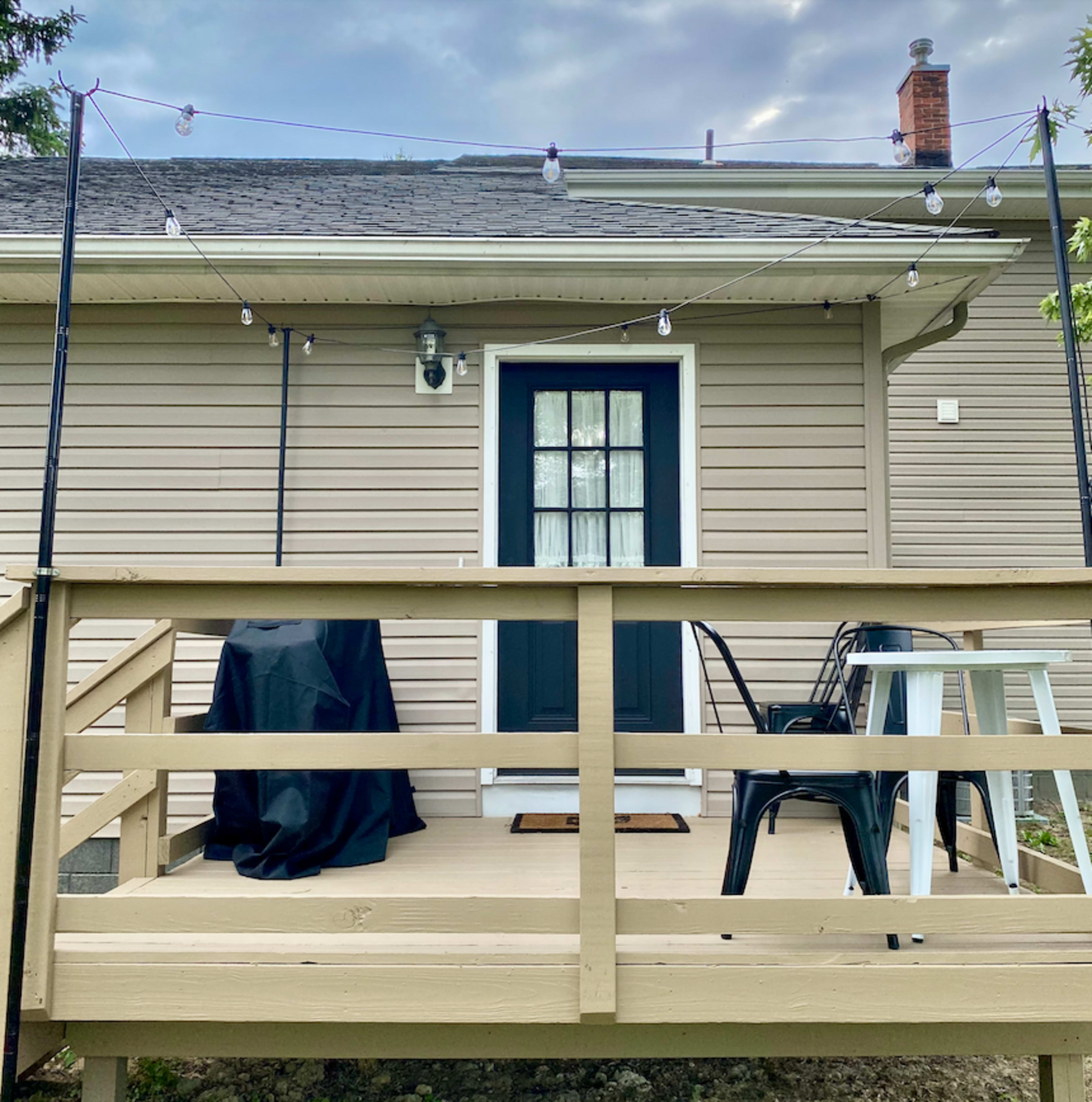 A wooden deck with two chairs, a table, and a covered grill, leading up to a door with a window.