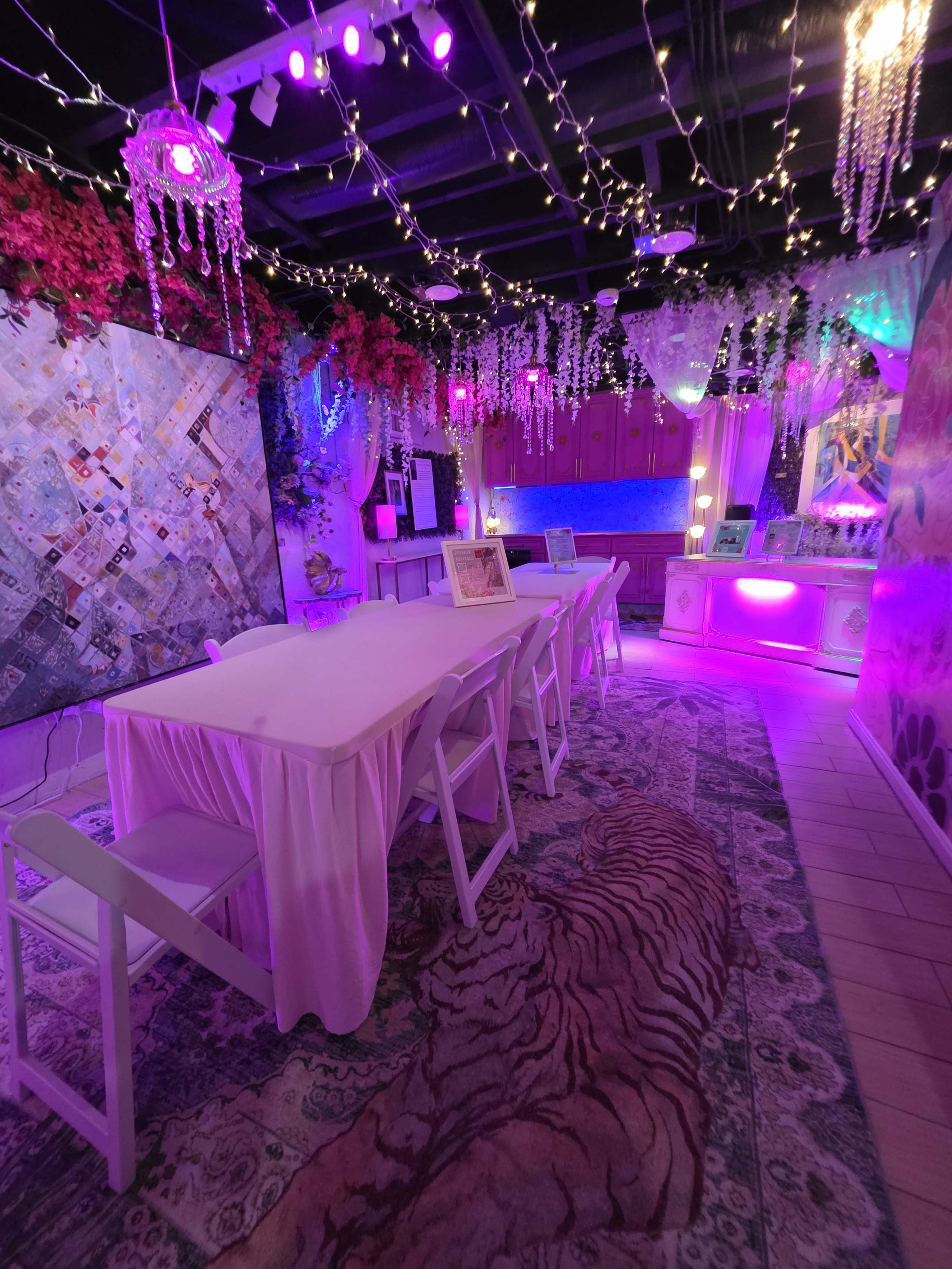 The image shows a brightly lit event space decorated with string lights, colorful artwork, and a long table covered with a white tablecloth.