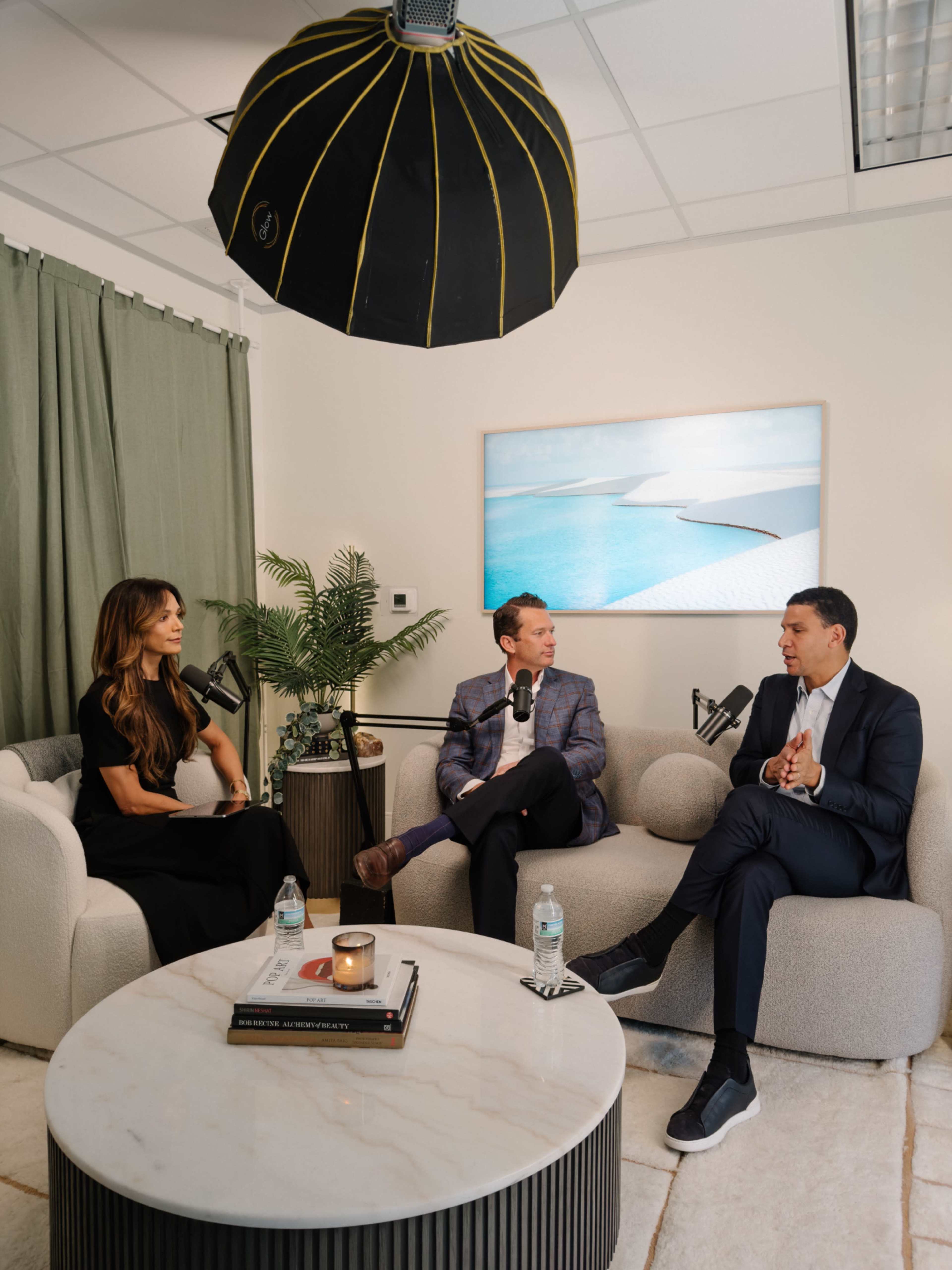 Three individuals are engaged in a discussion while sitting on a light-colored sofa in a modern, well-lit studio setting, with a large light fixture overhead and a beach-themed artwork on the wall.