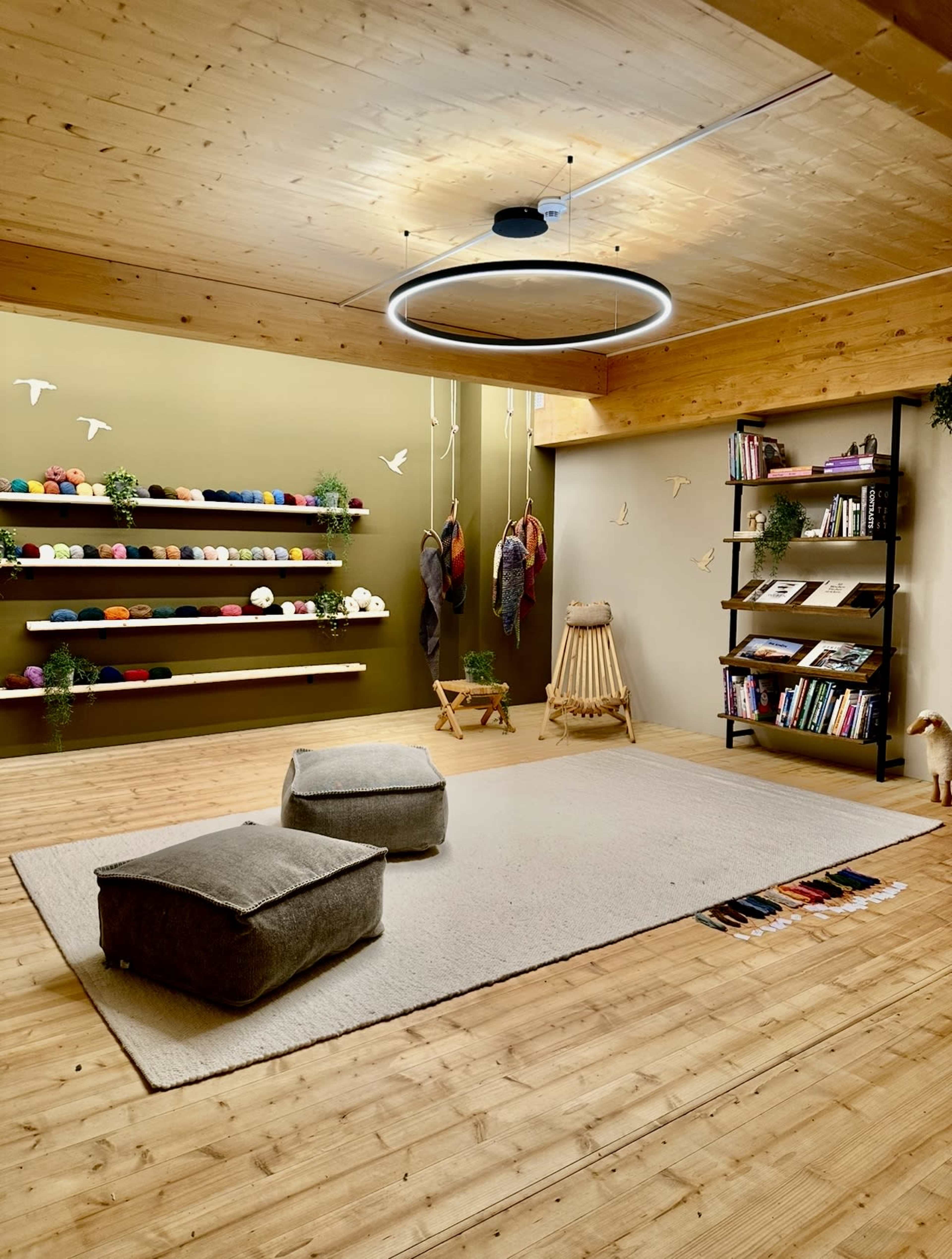 The image depicts a cozy, well-lit room with shelves displaying colorful yarn, a large rug on the wooden floor, and various seating options, including poufs and a wooden chair.