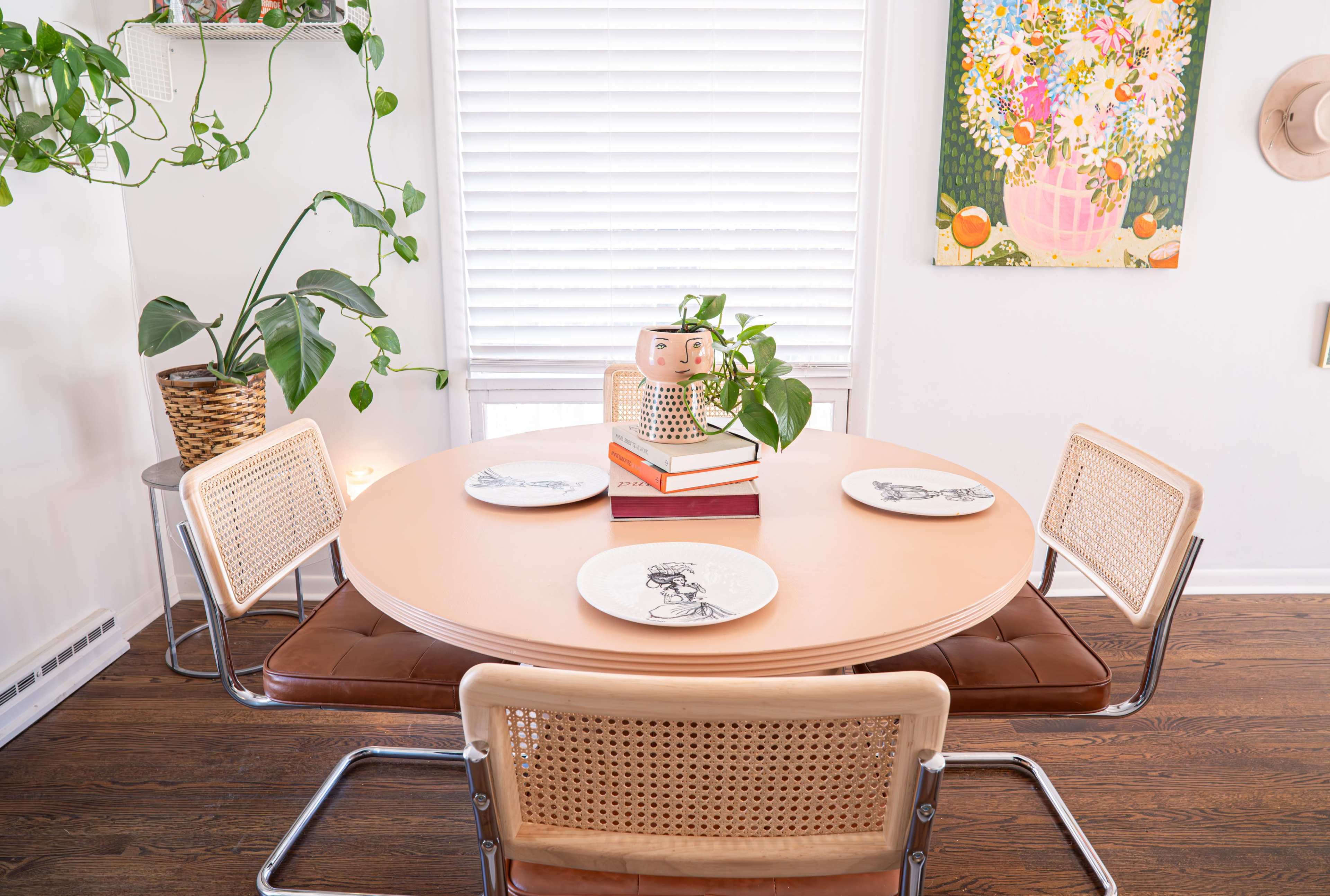 A round dining table with four chairs is set with decorative plates, a stack of books, and a potted plant, surrounded by plants and a colorful wall painting.