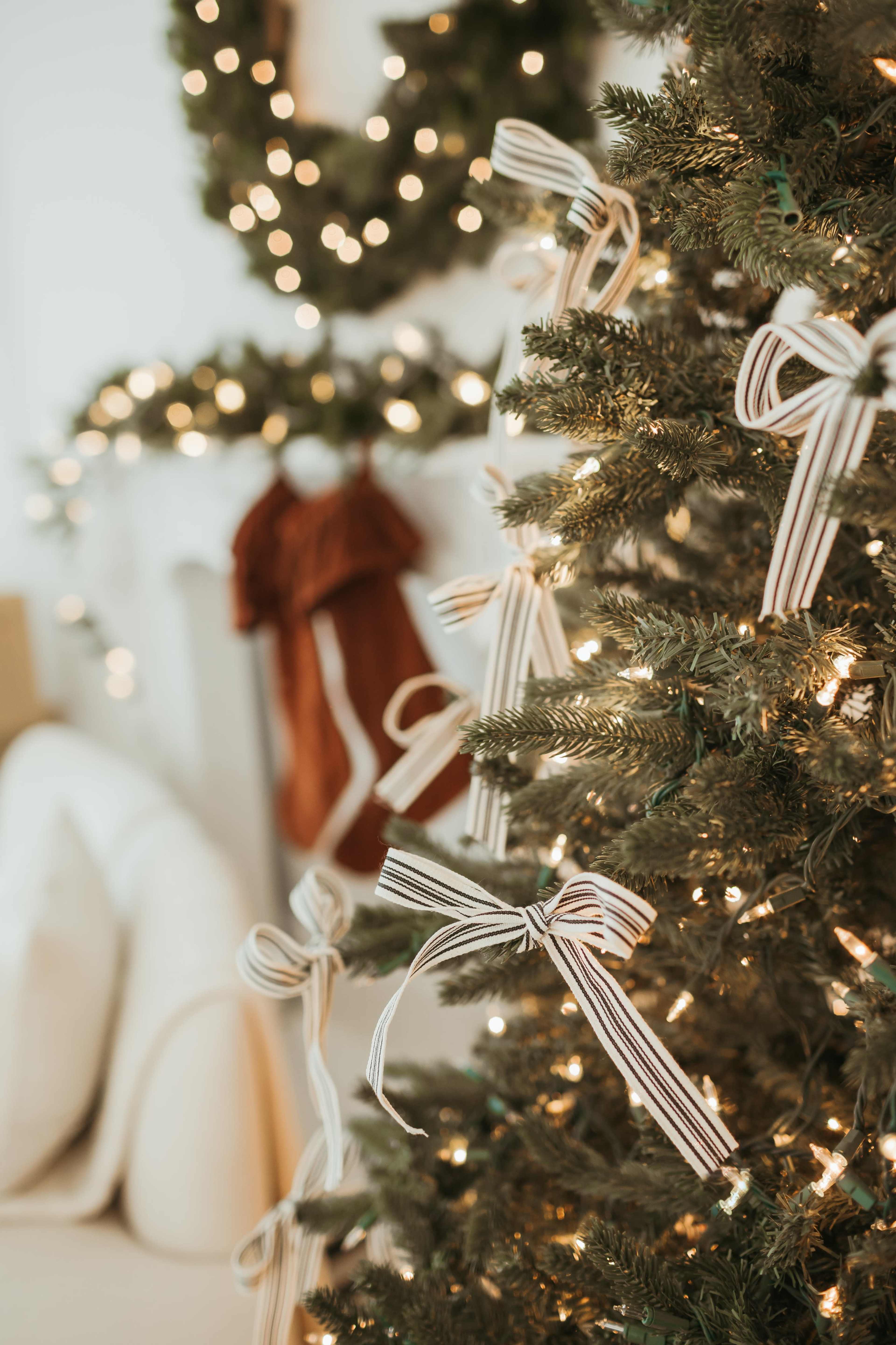 A decorated Christmas tree with striped bows and lights is positioned near a mantle adorned with greenery and stockings.