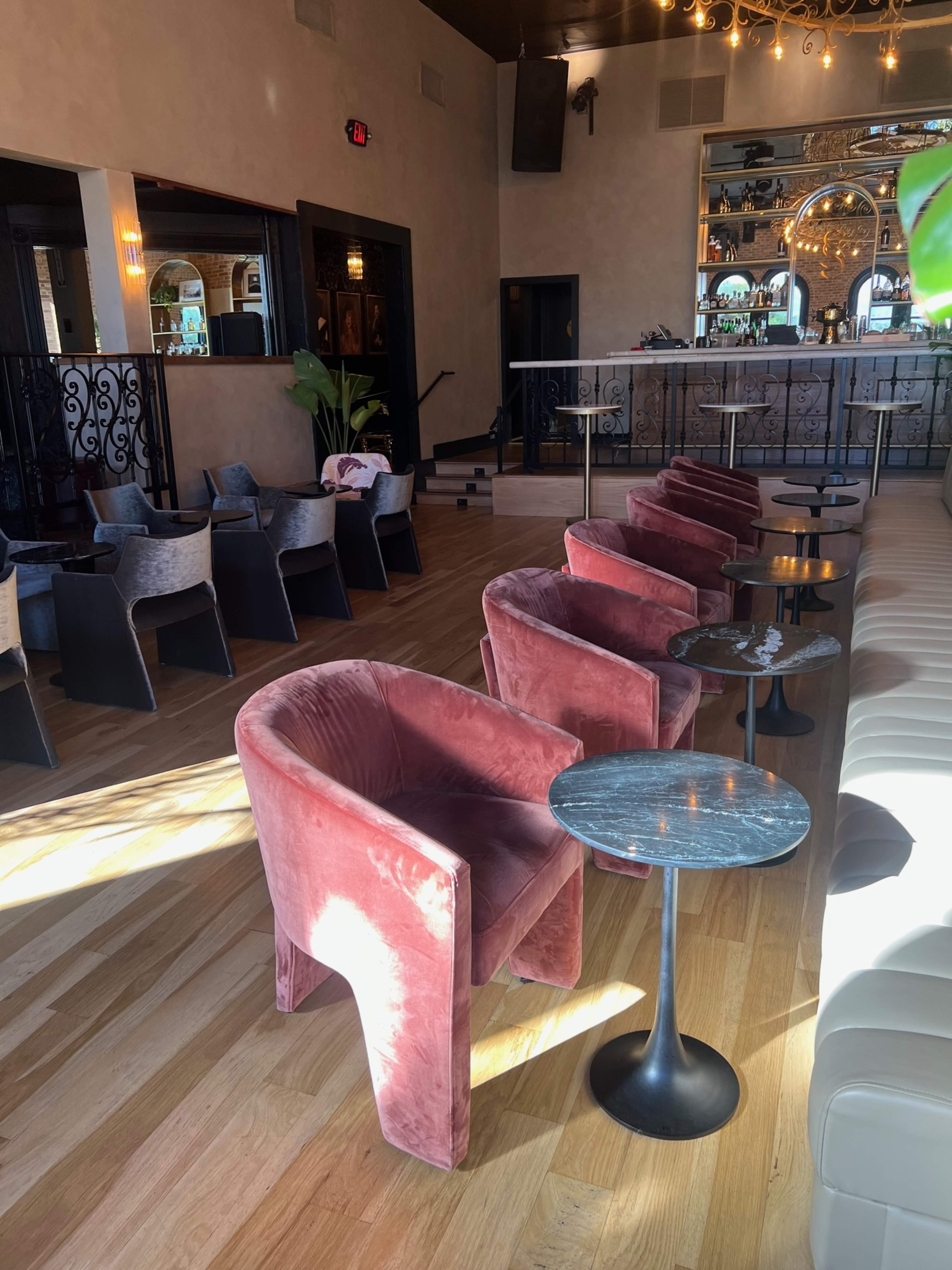 The image shows a lounge area with plush pink chairs and small black tables arranged across a wooden floor, with a bar visible in the background.