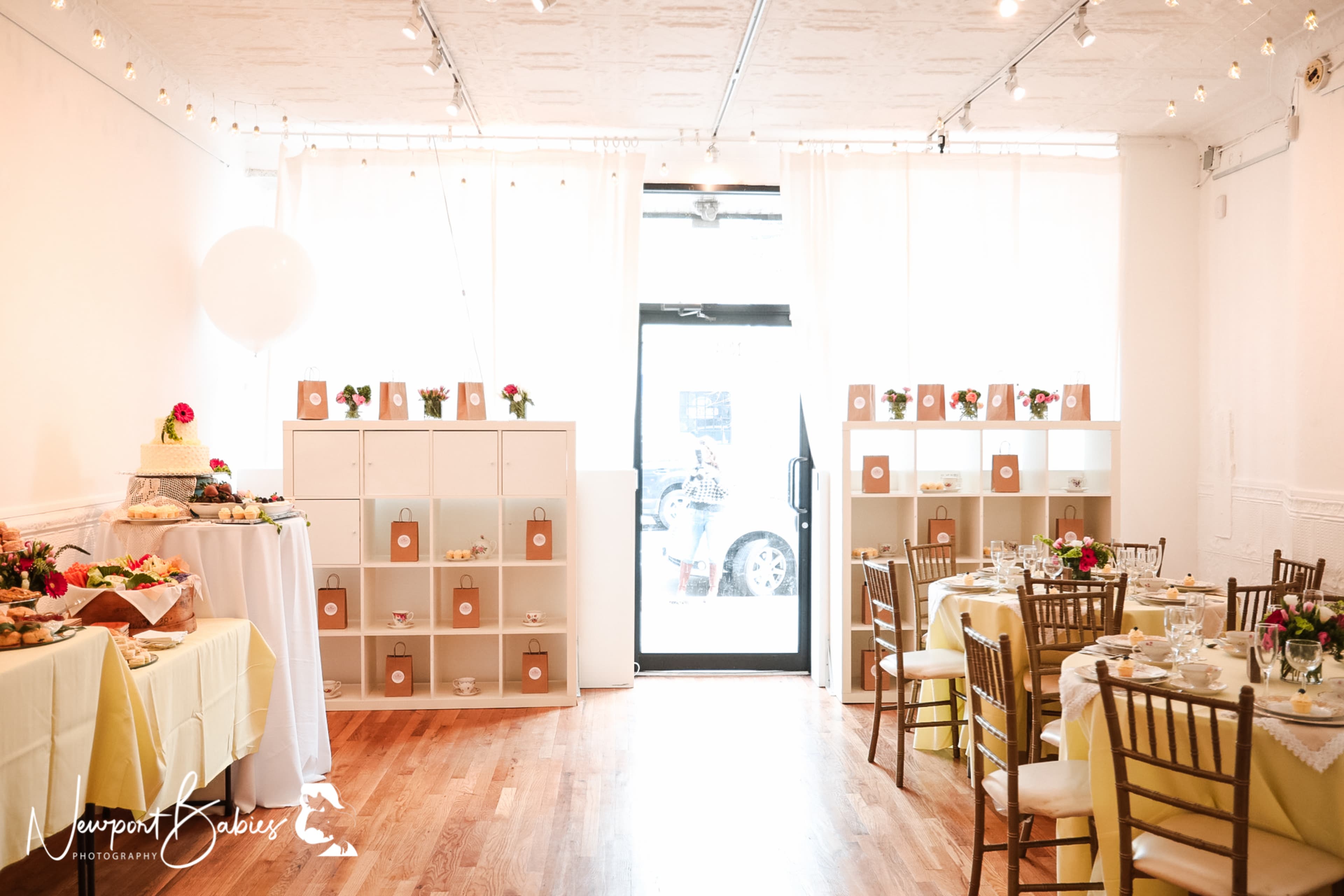 A bright, elegantly decorated event space with tables set for a gathering, featuring a food display on one side and a large entrance door on the other.