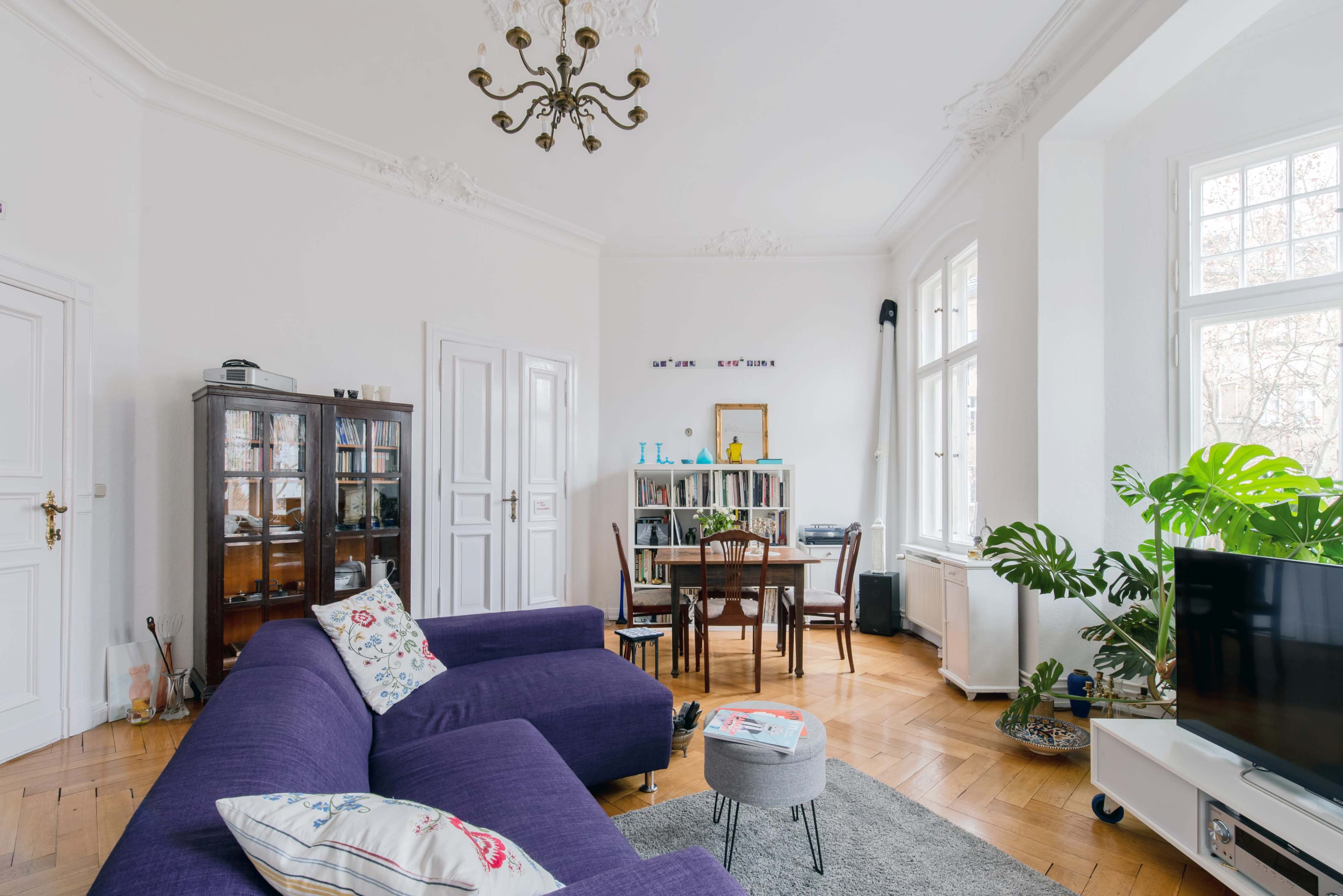 The image shows a bright living room with a purple sofa, a wooden dining table, and large windows allowing natural light to fill the space.