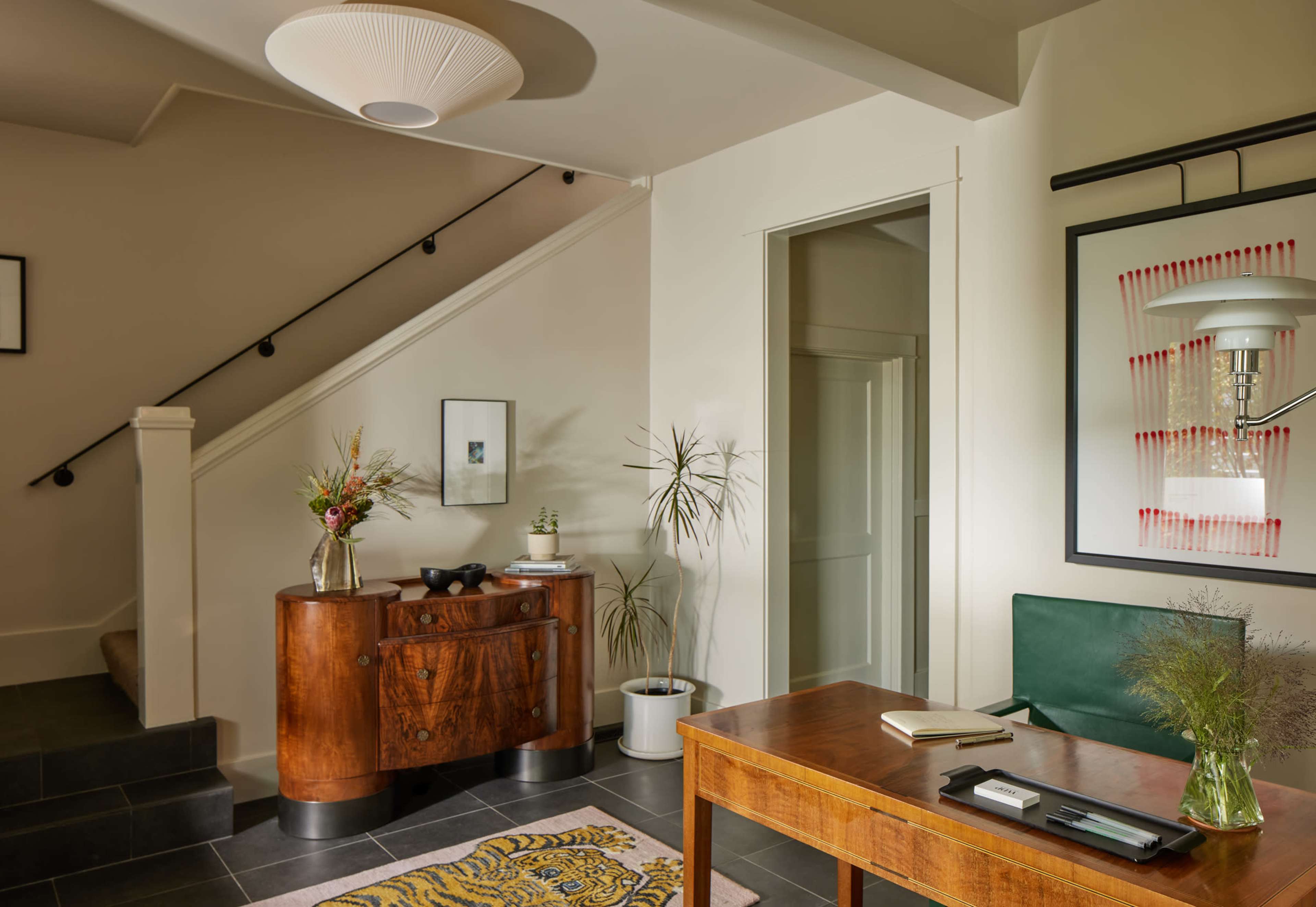 The image shows a modern entryway featuring a wooden desk, a green chair, a decorative cabinet, and a staircase in the background, with plants and artwork on the walls.